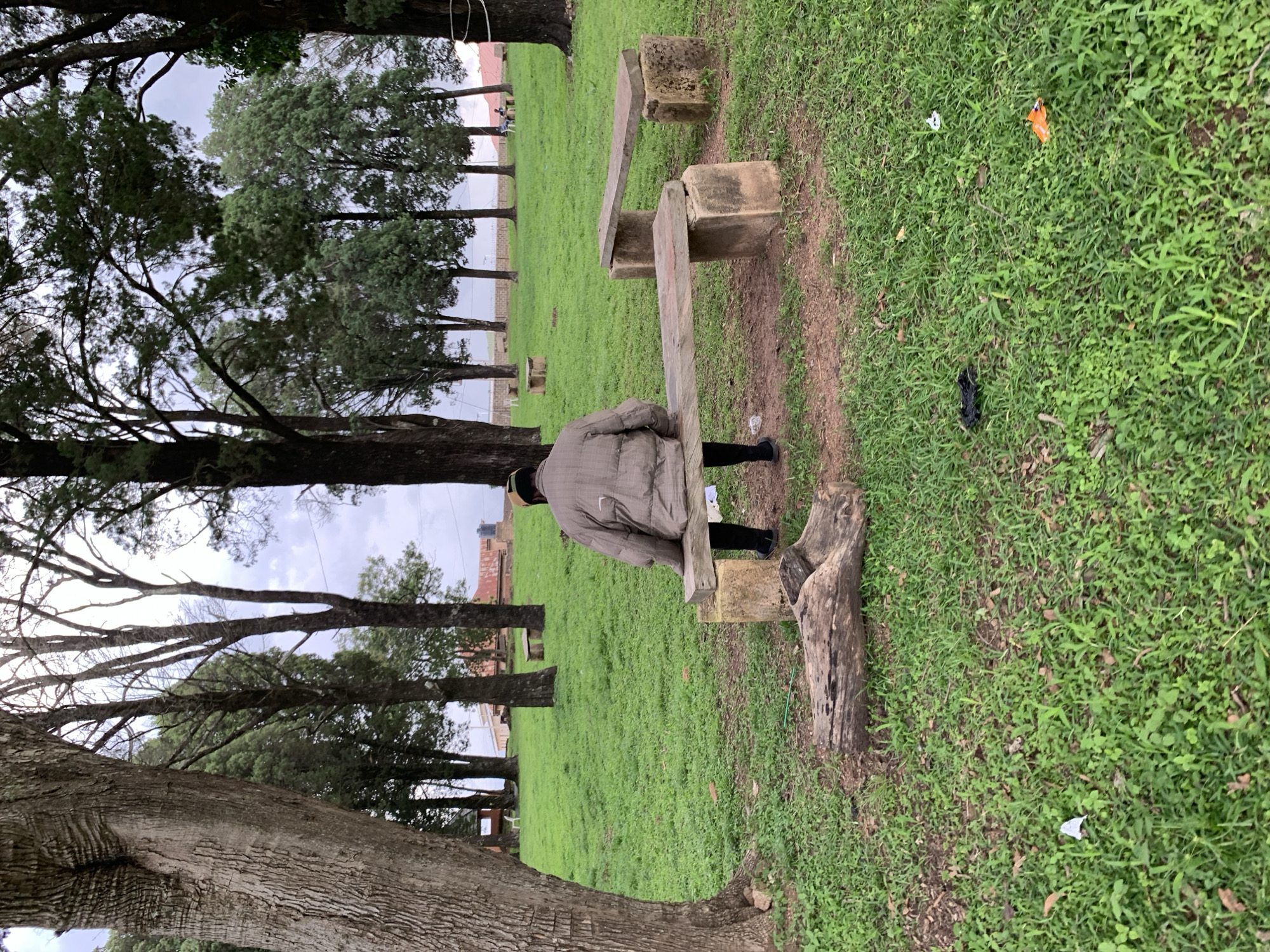 A person is sitting on a wooden bench supported by concrete blocks, facing away from the camera. The individual is wearing a brown quilted jacket and dark pants. The bench is situated in a grassy park area with several trees in the background. In the foreground, there are logs and scattered litter on the grass. The location is the recreational park in Jos, Nigeria. The sky is overcast.