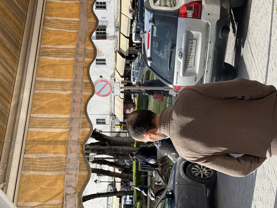 The image depicts an outdoor urban scene in the city of Tangier, Morocco, viewed from beneath a yellow and white striped awning. In the immediate foreground, the back of a male subject is visible, characterized by short, dark, thinning hair and a beard or stubble. He wears a brown, high-collared, long-sleeved sweater with his hands in his pockets, facing slightly left, away from the camera.
The midground features a paved street with vehicular traffic and pedestrians. A silver Kia Sportage SUV is prominently positioned on the right, displaying a Moroccan license plate reading "27742-أ-16". A dark grey car is partially visible on the left. Behind these vehicles, a white and red circular road sign indicates "STATIONNEMENT INTERDIT" (No Stopping/Parking). Several pedestrians are present; one individual in a dark jacket with a light-colored shoulder bag walks from left to right in the mid-distance. A motorcycle or scooter is also visible on the left side of the street.
The background reveals a public square or park area with numerous mature trees, currently bare of leaves. Beyond the trees, white multi-story buildings with numerous rectangular windows, some fitted with brown shutters, line the far side of the square. The ground levels of these buildings house shops, each featuring a beige fabric awning. The scene is illuminated by strong sunlight, casting distinct shadows.