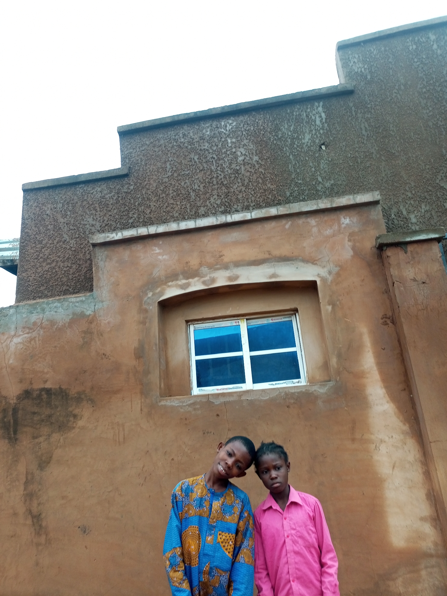 In the city of Tempure, Nigeria, two children, a boy and a girl, stand against a textured, ochre-colored wall. The boy, on the left, wears a vibrant blue and yellow patterned tunic, his head tilted towards the girl with a smile. The girl, on the right, is dressed in a bright pink collared shirt, her expression neutral as she looks towards the camera. Behind them, a window with white framing and blue panes is set into the wall. The building's facade is weathered, with visible cracks and darker splotches indicating age and exposure to the elements. Above the window, the building's architecture features stepped sections, with a rougher, darker textured material on the upper levels. The sky above is a bright, overcast white.