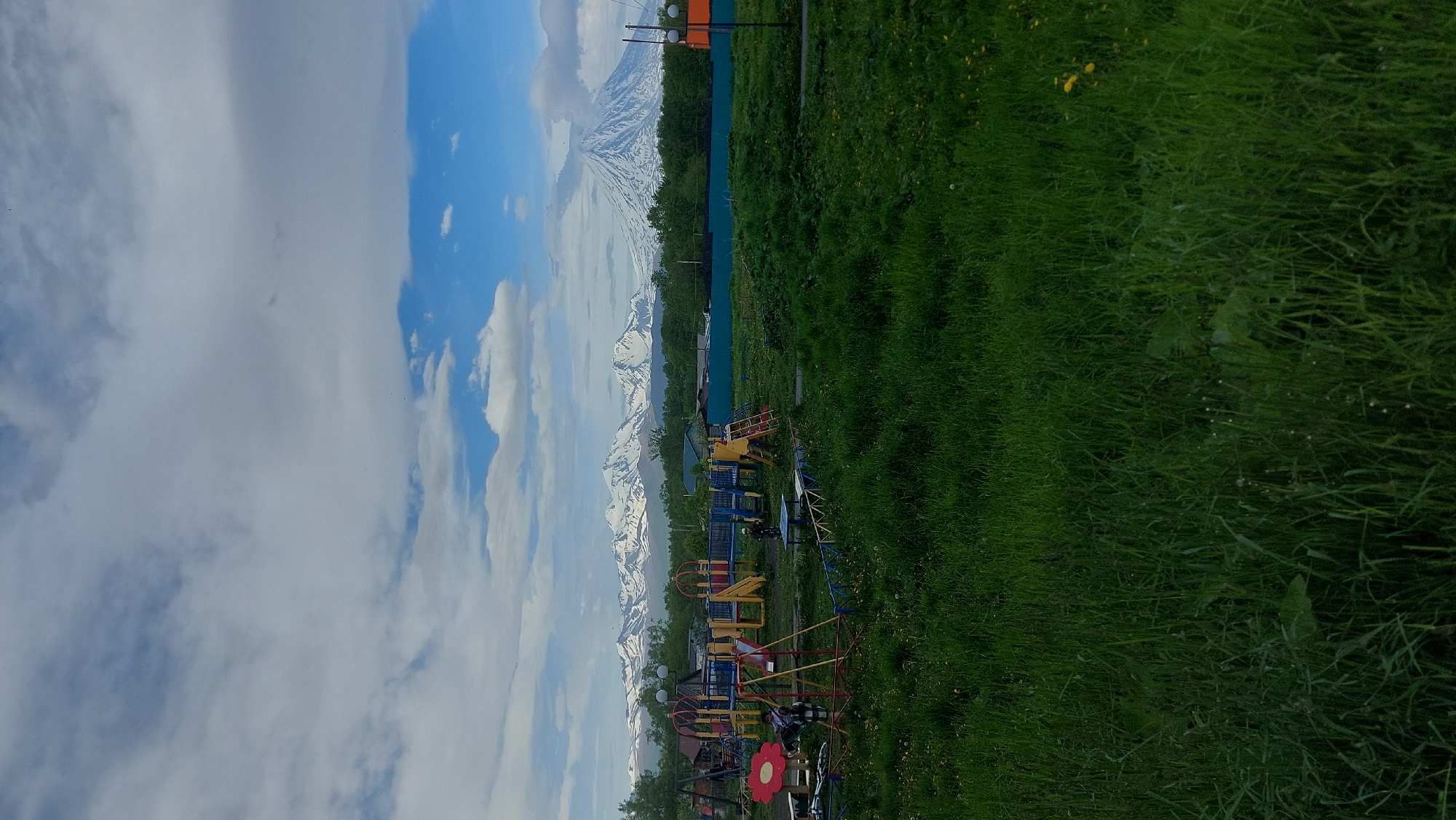 The image depicts a playground in the foreground, featuring a red flower-shaped swing seat, a metal swing set with yellow and red poles, a blue and yellow slide structure with a ladder, and other playground equipment. Two individuals are visible on the playground, one near the swing and another standing by a small table. The playground is surrounded by a low blue and white fence, and the ground is covered in green grass with scattered yellow flowers. In the middle ground, there are several residential buildings with blue roofs, and a teal-colored fence runs horizontally across the scene. Behind the buildings and fence, a dense line of green trees is visible. The background is dominated by a majestic mountain range with snow-capped peaks, and a prominent conical mountain with a snow-covered peak and a plume of smoke or cloud emanating from its summit. The sky above is partly cloudy with patches of blue. The overall scene suggests a rural or semi-rural setting in Зелёный, Russia, with a playground in the foreground and imposing volcanic mountains in the distance under a dynamic sky.