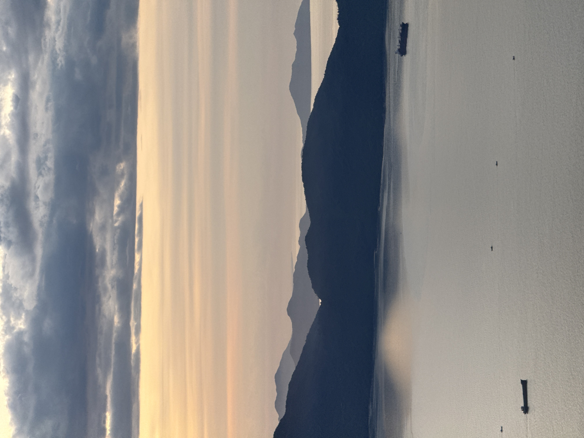 The image depicts a wide, tranquil bay at dusk, with layered mountain ranges receding into the distance. The sky is a gradient of soft orange and yellow near the horizon, transitioning to shades of gray and dark blue as it reaches the top of the frame, with a dense layer of clouds. The water is calm and reflects the muted colors of the sky and the dark silhouettes of the mountains. Several small boats are visible on the water, along with a larger cargo ship in the middle ground. The scene is serene and evokes a sense of natural beauty and peacefulness. This scene is likely the Son Tra Peninsula or the waters surrounding it in Da Nang, Vietnam, a popular location for its scenic coastal views and mountainous landscape.