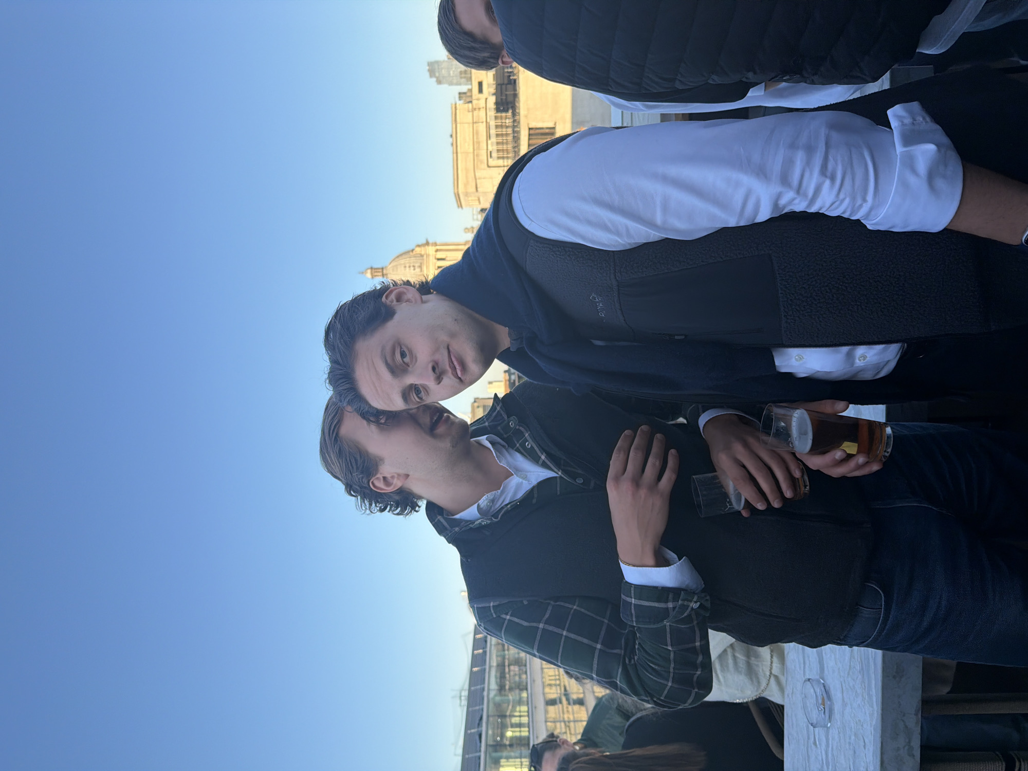The image shows a group of people outdoors, likely on a rooftop or high terrace, as indicated by the cityscape visible in the background. Two men are prominently featured: one is wearing a white shirt and a dark vest and another is wearing a green plaid shirt and a dark vest; both are holding drinks. The cityscape includes buildings recognizable as being in London, United Kingdom, most notably a domed building similar to St. Paul's Cathedral. Other individuals are partially visible around the edges of the image. The overall atmosphere suggests a casual gathering or social event.