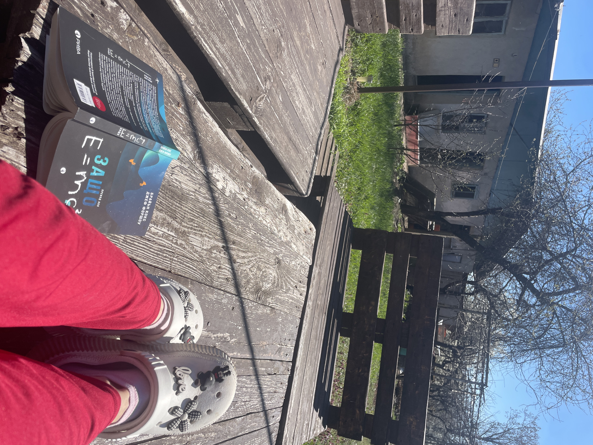 The image shows a person sitting on a weathered wooden deck in what appears to be an outdoor space. The person is wearing red pants and white clogs adorned with charms. Open on the deck is a book titled "E=mc²?" by Brian Cox and Jeff Forshaw in Bulgarian translation. The book is published by RIVA. Behind the deck, there is a patch of green grass, a building with broken windows, and bare trees against a clear blue sky. The location is not clear enough to ascertain it is in Sofia, Bulgaria.