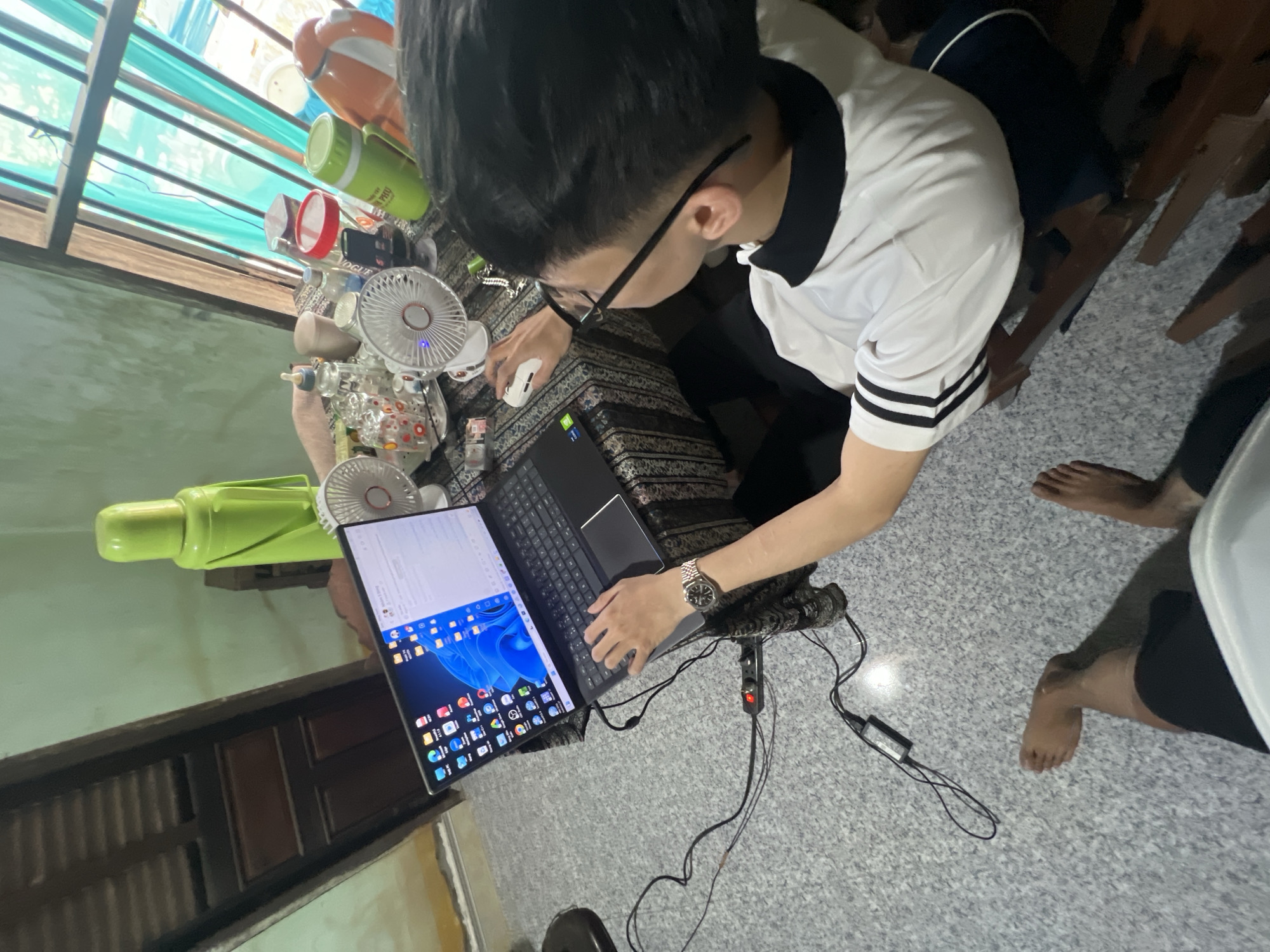 This image depicts an indoor scene in Bình An, Vietnam, showing an individual engrossed in computer work. The main subject, a person with short dark hair and glasses, wearing a white and black polo shirt, is viewed from an overhead perspective. They are seated on the floor or a low surface, intently focused on a black laptop and an external monitor positioned on a patterned fabric, possibly a blanket or mat. Their left hand manipulates a white computer mouse, while their right hand rests on the laptop's keyboard. A silver watch is visible on their left wrist.
The workspace is adorned with several personal items, suggesting a casual, domestic environment. To the left of the laptop, at least three small portable fans are visible, indicative of warm weather. Various containers, including an orange thermos, a red-lidded jar, and a plastic bottle, are scattered on the surface. A green insulated container bears the Vietnamese text "PHƯƠNG," and a small object displays the word "VOGUE." A power strip with a red indicator light lies on the grey speckled floor, connected to several cables.
The laptop's external monitor displays a desktop interface with a blue abstract wallpaper and various application icons, characteristic of a Windows operating system. The laptop's own screen shows text-based content, potentially code, a document, or a webpage. In the background, windows covered with translucent green/blue sheeting filter natural light, and a dark wooden door or partition is partially visible. The bare feet and lower legs of another person are visible on the right side of the frame, suggesting shared space. The overall scene portrays a focused work or study session in a comfortable, informal setting, likely during the daytime.