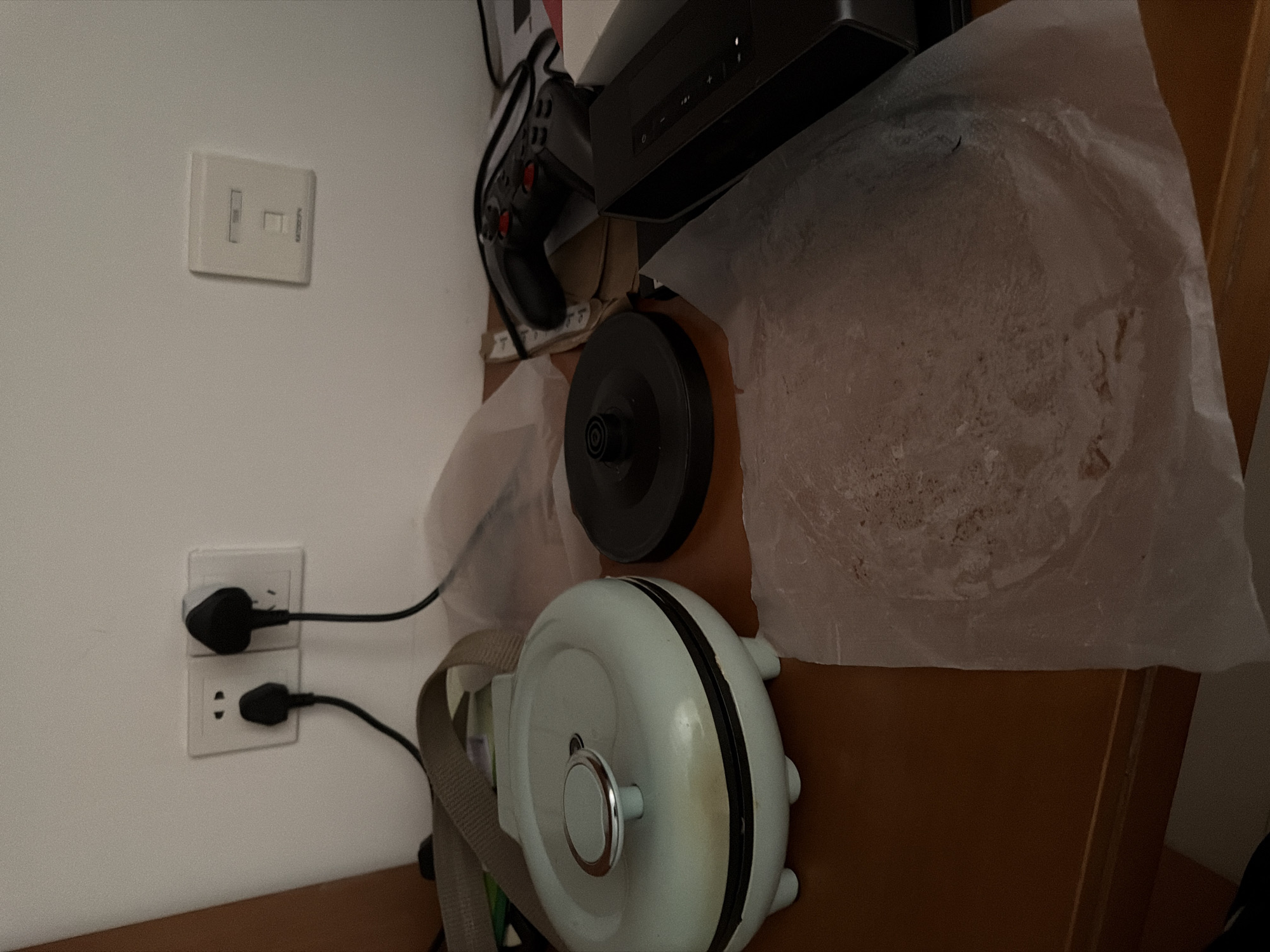 The scene depicts a cluttered domestic environment. An electrical outlet on a white wall has two plugs inserted. Adjacent to the outlet is a light switch. A small, round, light blue appliance with a carrying strap sits on a wooden surface. A black kettle base is next to it, partially covered by a piece of wrinkled, translucent paper or plastic. Further back is a black gaming controller and a black electronic device, possibly a streaming device. The background is indistinct. The city of Jiangning District, China, cannot be definitively determined from the image.