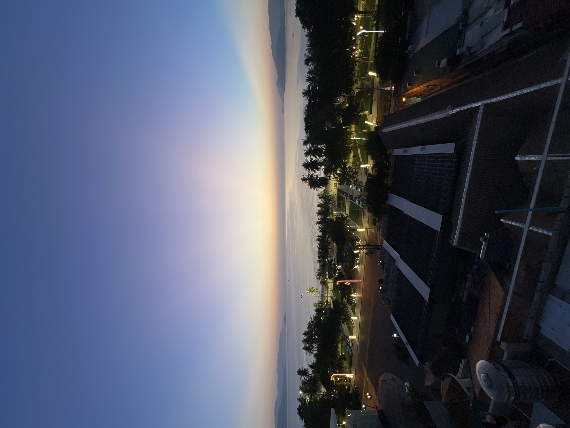 The image captures a twilight view of Nha Trang, Vietnam, overlooking the coastline. The sky transitions from a deep blue at the top to softer hues of purple, pink, and orange near the horizon, indicating either dawn or dusk. A calm sea stretches towards distant, hazy mountains. Along the coast, a line of palm trees is silhouetted against the lighter sky. In the foreground, illuminated streetlights line a road and a park area, casting a warm glow on the lush greenery and paved pathways. Beyond the park, several buildings with dark roofs are visible, some featuring lighter-colored paneling. The overall scene depicts a tranquil urban coastal environment as day transitions into night.