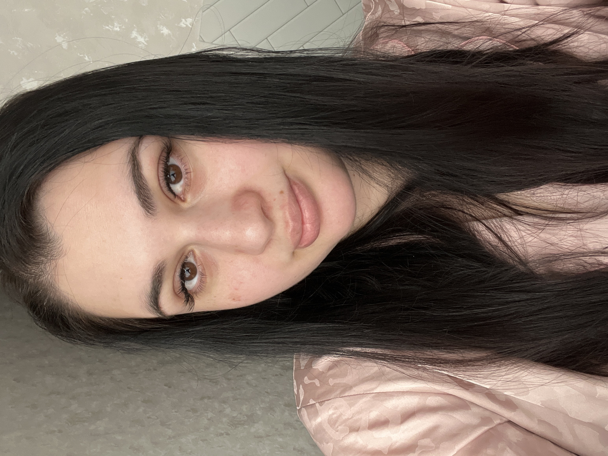 A close-up, horizontally oriented shot depicts a young woman with long, straight, dark brown hair. She lies with her head tilted, gazing directly at the camera with a gentle, closed-mouth smile. Her skin is fair, and she has dark brown eyes, well-groomed eyebrows, and visible mascara on her eyelashes. A faint reddish mark is present below her right eye, and a small mole is visible above her upper lip on the right side of her face.
She is wearing a light pink garment, possibly a robe or sleepwear, made of a silky or satin-like fabric with a subtle, lighter-toned patterned texture. Her hair cascades around her face and extends towards the bottom and right side of the frame, partially resting on the pink fabric of her clothing.
The background behind her head is a light-colored tiled surface, possibly a wall, while the area to the right of her hair and body shows more of the pink patterned fabric, suggesting she might be lying on a bed or couch. The lighting is soft and even, indicating an indoor setting. No specific time of day or weather conditions are discernible from the image. The overall mood is relaxed and calm. No visible text is present. The context provided indicates the location is Chaikovsky, Russia.