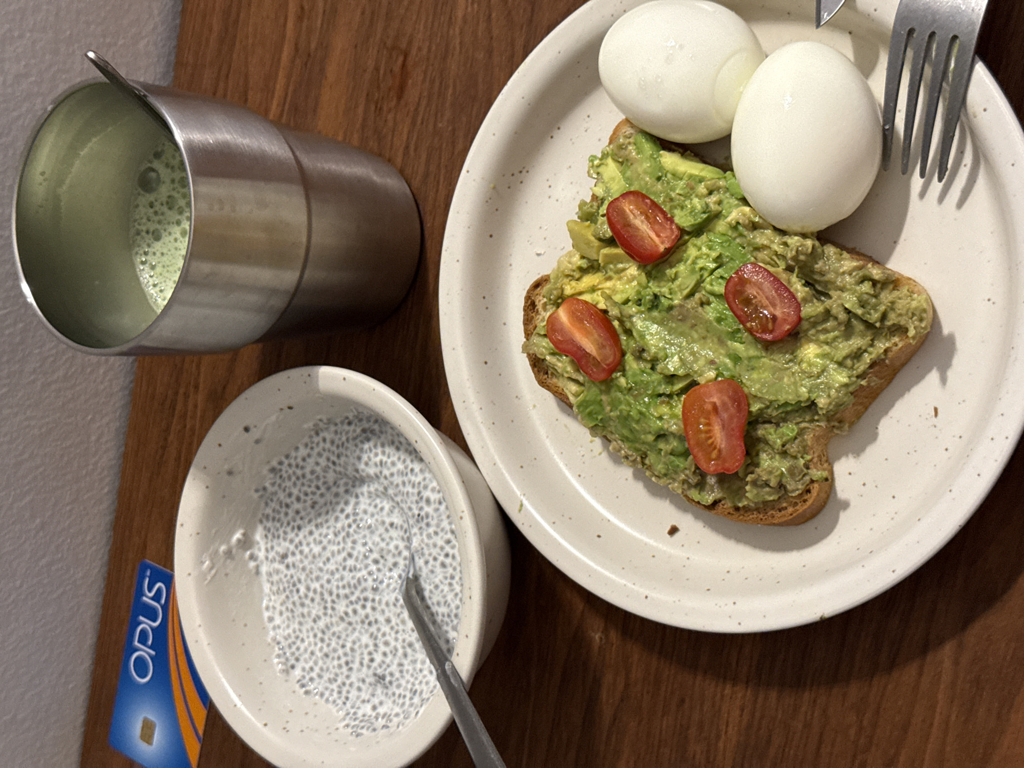 A top-down shot captures a breakfast or brunch meal arranged on a warm, wooden surface. The main focus is a light-colored, speckled plate holding a slice of avocado toast topped with mashed avocado and three halved cherry tomatoes. Two whole, peeled boiled eggs sit beside the toast, with a silver fork resting on the plate's upper right edge.
To the left of the plate, a small, light-colored bowl contains chia pudding, characterized by numerous black seeds suspended in a white liquid, with a silver spoon immersed in it. A blue and orange card, partially visible next to the bowl, displays the word "Opus," indicating a location in Montreal, Canada, as this is its public transport fare card.
In the upper left corner, a brushed metal tumbler contains a frothy, light green beverage, likely matcha, with a small silver stirrer or spoon inside. The items are neatly arranged, suggesting a prepared meal ready for consumption in an indoor setting. The lighting is warm and even.