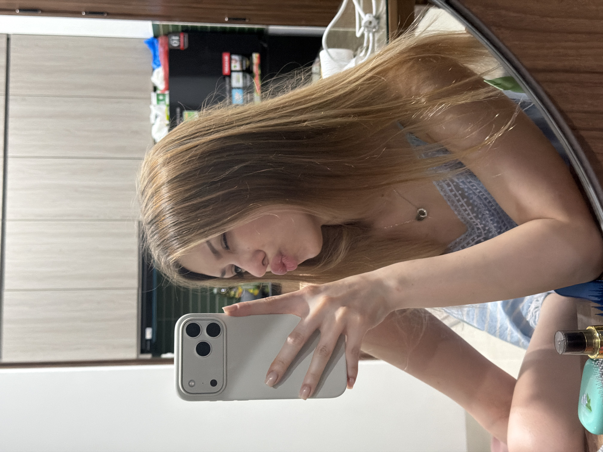 A young woman is taking a selfie in a mirror. She is holding a white iPhone with three cameras on the back. She is making a kissing face. She has long blonde hair. She is wearing a blue and white top. A silver necklace with a charm is visible on her neck. The background shows a wooden cabinet and a white wall. A small perfume bottle and hairbrush are to the bottom right. The location is indeterminable as there is no information to determine if this photograph was taken in Đà Nẵng, Vietnam.