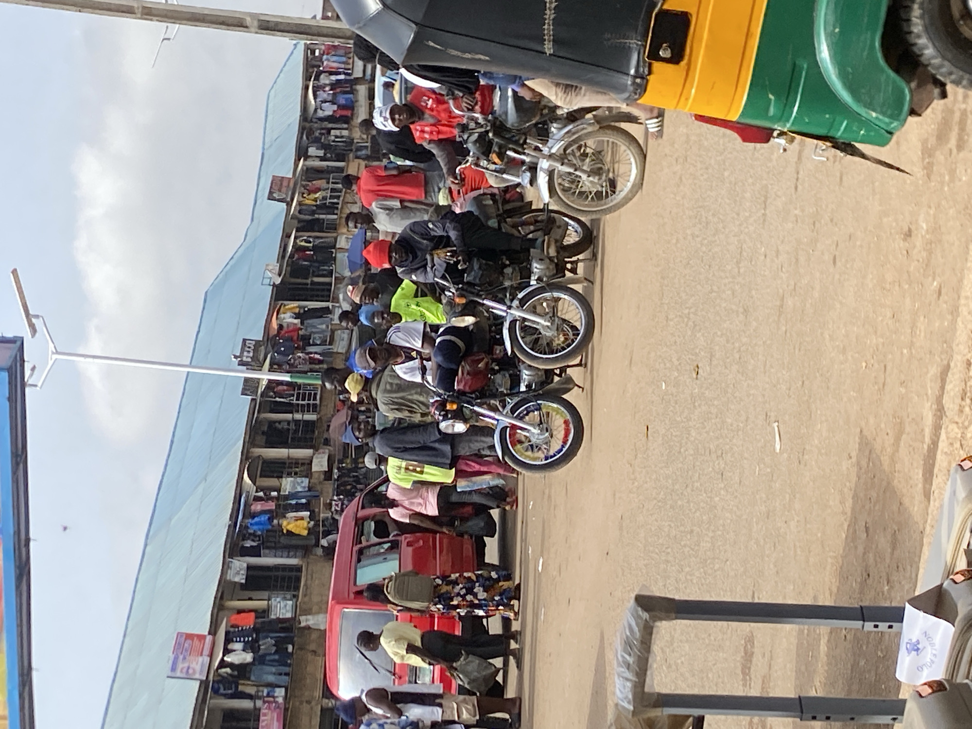 The image captures a bustling street scene in Bukuru, Nigeria. Several motorcycles are lined up along the road, with riders seated on them. A three-wheeled vehicle, painted in green, yellow, and black, is partially visible at the top of the frame. Alongside the road, a multi-story building houses shops or stalls. Pedestrians are visible walking along the sidewalk. The sky is overcast.