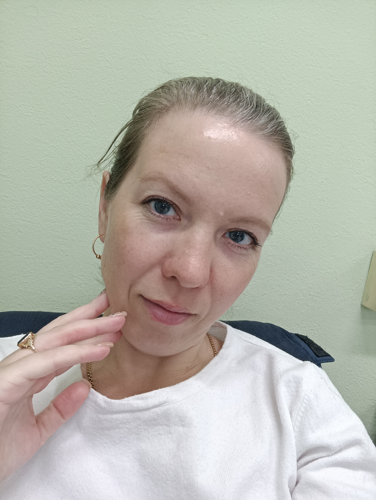 A close-up selfie of a middle-aged woman with fair skin, blue eyes, and light gray hair pulled back. She is wearing a white t-shirt and has a gold necklace and hoop earrings. She is wearing a ring on her right hand and that hand is touching her face. The background is a light green wall. There is no definitive evidence to place the location in Chaikovsky, Russia.