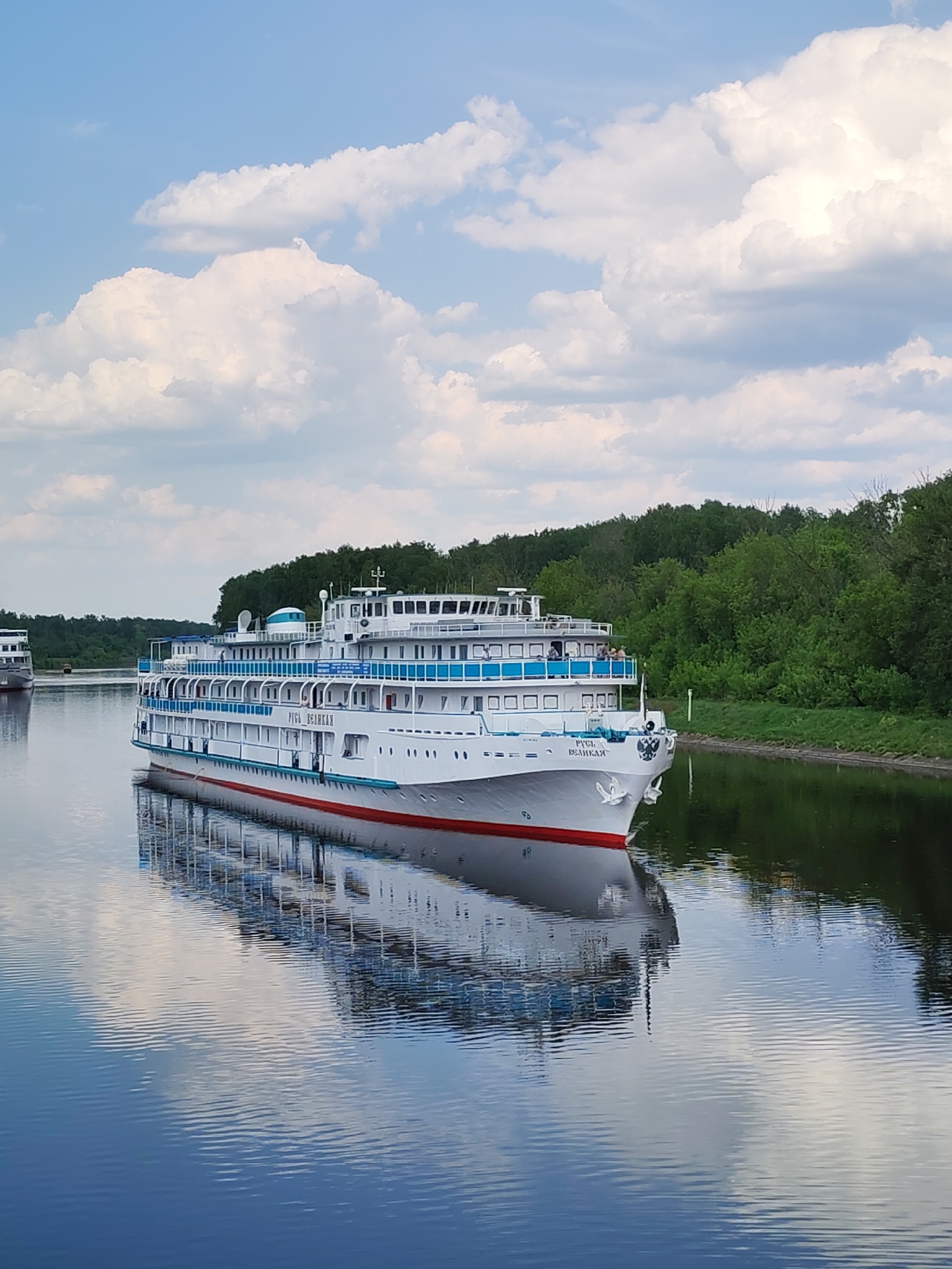 The image depicts the river cruise ship "РУСЬ ВЕЛИКАЯ" (Rus Velikaya) sailing on a waterway near Yakhroma, Russia. The ship is white with blue railings and a red waterline stripe. The name of the ship is printed in Russian on the bow. A reflection of the ship and the sky is visible on the calm water surface. The surrounding landscape consists of green trees and bushes on the banks of the waterway. The sky is blue with scattered white clouds. Another ship is visible in the distance.