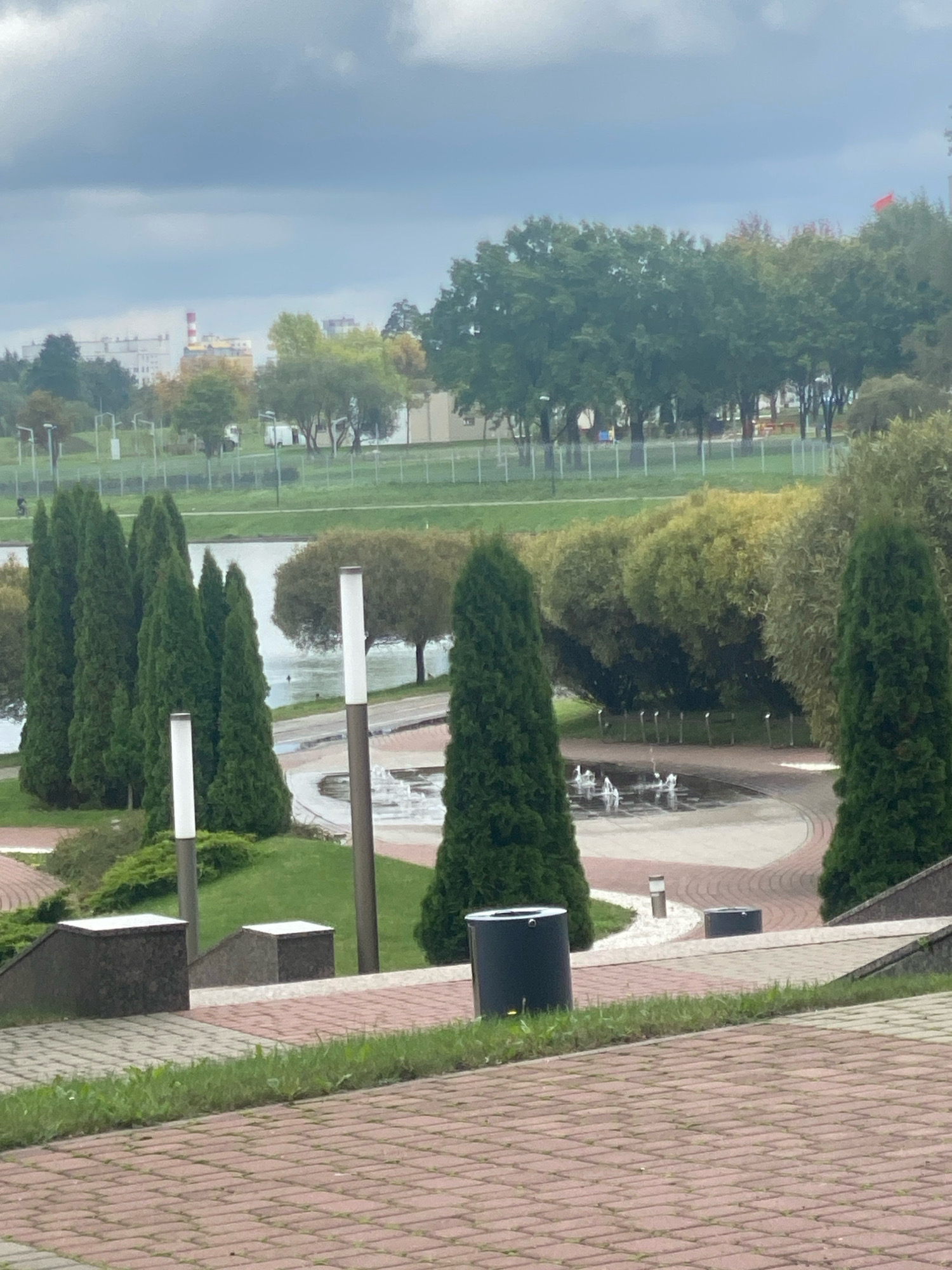 The image captures a section of an urban park in Minsk, Belarus, under an overcast sky. In the foreground, a paved pathway made of reddish-brown bricks leads the eye upwards. To the left of the pathway are two dark gray cylindrical trash receptacles. A low-lying green lawn borders the pathway.
Further into the scene, a series of dark gray planters with white tops line a tiered pathway, interspersed with green grass and small bushes. A cluster of tall, conical evergreen trees dominates the mid-ground, with several more on either side. Beyond the trees, a body of water, likely a river or lake, is visible.
In the background, a circular area features a series of small water fountains actively spraying water. A paved area surrounds the fountains, with curved pathways. Beyond the water feature, a row of trees with varied foliage, some appearing autumnal in color, stretches across the landscape. In the far distance, buildings and a red and white smokestack are visible, along with a red flag on a tall pole. The overall impression is of a park setting with recreational amenities, bordering a natural water body, within a city.