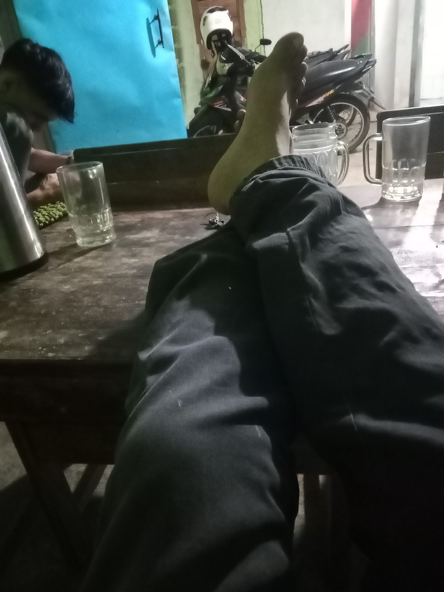 In a dimly lit room, a person's legs, clad in dark pants, are prominently displayed, crossed over a wooden table. The camera angle is low, focusing on the lower half of the body. To the left, a person with dark hair is partially visible, leaning forward, with their hands on the table, near a glass of liquid and what appears to be a pile of green beans or peas. A metallic thermos-like object is also on the table. To the right, two more glasses are present, one with a handle. In the background, a motorcycle with a white helmet on it is visible. The environment suggests a casual setting, possibly a home or a small eatery in Leksono, Indonesia, given the presence of a motorcycle and the general ambiance. The lighting creates distinct shadows, adding to the overall mood of the scene.
