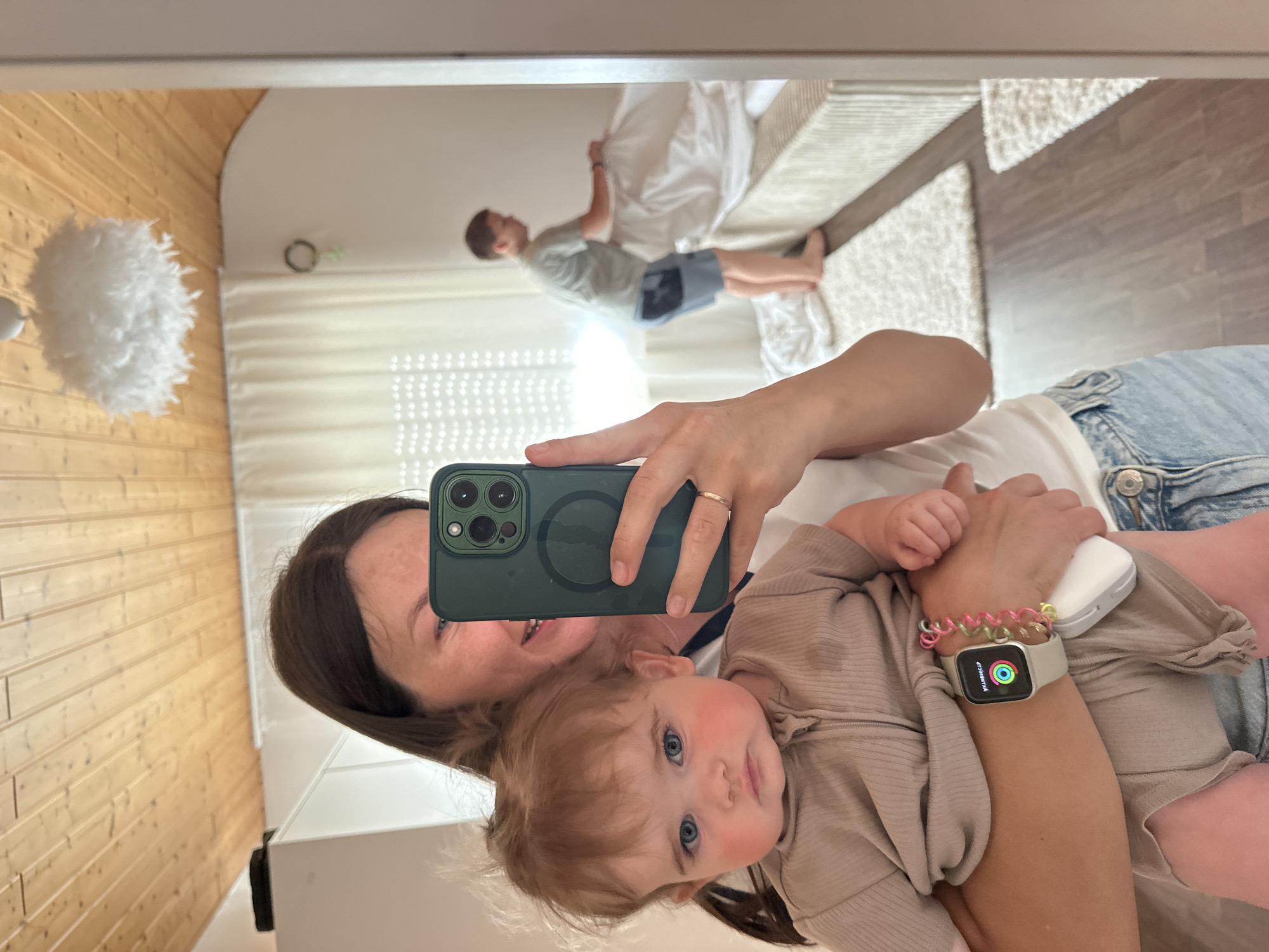 A woman holding a baby is taking a selfie in a mirror. The reflection shows a room with wooden walls, a white feather lamp, and a boy on a bed. The woman is wearing a white shirt and jeans. The baby is wearing a tan outfit. The woman is holding a green phone with a magnetic charger attached. She is also wearing a silver Apple Watch with a colorful band. The location is indoors, likely a home. There is no specific evidence to place this in the city of Niederquembach, Germany.