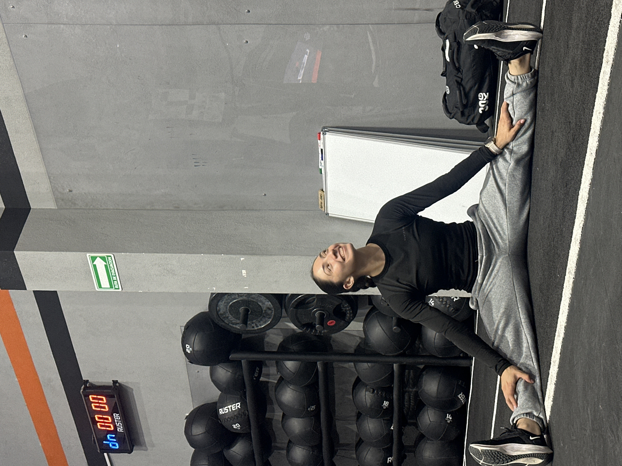 The image depicts a woman in a gym setting in San Bernardino Tlaxcalancingo, Mexico. She is performing a split, with one leg extended along the wall and the other leg extended in the opposite direction, resting against the edge of the flooring. The woman is wearing a long-sleeved black shirt and grey sweatpants, with black and white running shoes. She is smiling and looking upwards.
Behind her is a rack filled with black medicine balls, each labeled with weights such as "20" or "14". To the left, there is a wall with orange and black stripes, and a digital timer displays "00 00". In the background, a whiteboard is visible. A black bag is partially visible near the wall.