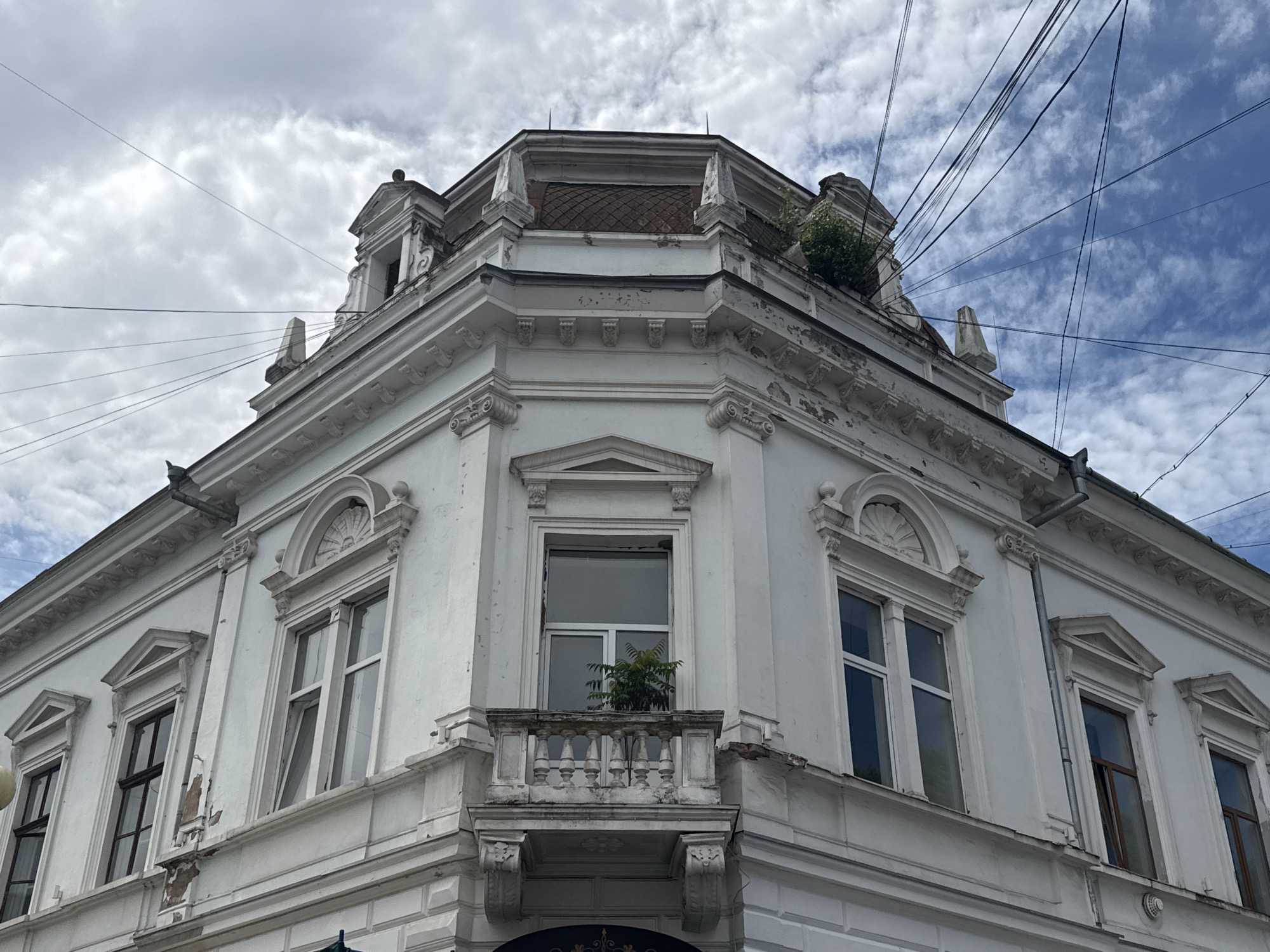 The image shows a corner of a white, multi-story building with ornate architectural details, located in Kolomyia, Ukraine. The building features multiple windows, some with decorative pediments above them, and a small balcony with a balustrade. The upper part of the building has a domed roof section with visible tiling and some greenery growing on the rooftop. A complex network of electrical wires crisscrosses the sky above the building. The sky is partly cloudy with patches of blue sky visible. The paint on the building shows signs of wear and peeling in places. The photograph is taken from a low angle, looking upwards at the building.