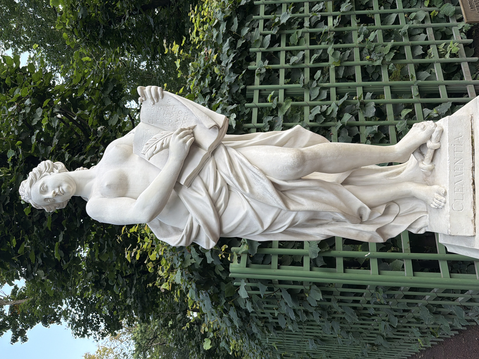 The image shows a white marble statue of a female figure, likely allegorical, standing outdoors. The statue is draped in flowing robes and holds an open book in her hands, with a quill pen resting on the page. The text on the book appears to be in Latin and includes the word "CLEMENTIA" which is also inscribed on the pedestal of the statue. The statue is positioned in front of a green trellis fence covered in ivy, with lush trees and foliage in the background under a clear sky. This sculpture is Clementia, located in the Garden of the Hermitage in the State Hermitage Museum, Saint Petersburg, Russia.