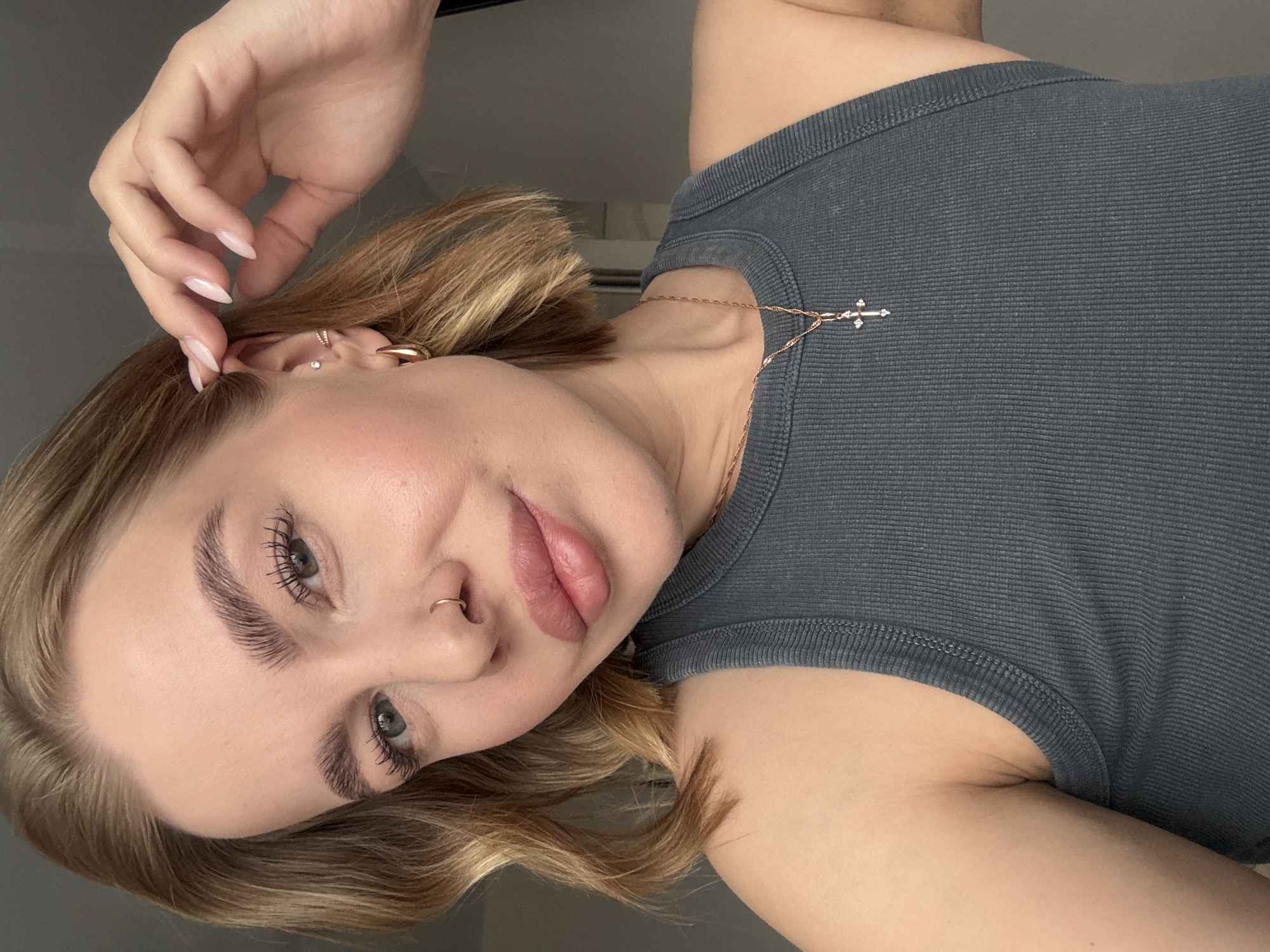 A woman is centered in the frame, angled slightly to the left. She has light skin, blonde hair, and green eyes. Her lips are full, and she has a nose ring in her left nostril. She is wearing a gray ribbed tank top and a gold necklace with a cross pendant. Her left hand is raised near her head, with long, pointed nails. She also has multiple earrings in her left ear. The background is blurred, suggesting a room with a window. There is no evidence to suggest the location in the city of Chaikovsky, Russia.