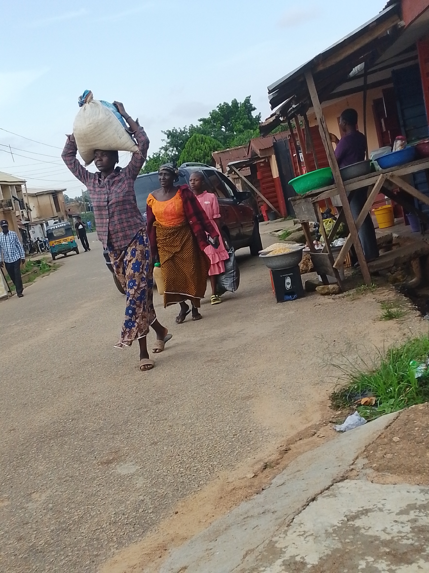 The image shows a street scene in Jos, Nigeria, likely in the afternoon given the lighting. Several people are walking along the paved road. In the foreground, a person wearing a plaid shirt and a patterned skirt carries a large sack of goods on their head. They are walking alongside another person in an orange top and brown patterned skirt, and a third person in a pink dress. The background reveals a street with buildings and market stalls. A car and a three-wheeled taxi are also visible on the road. One stall displays various bowls and containers, some filled with what appears to be grains or produce. The overall atmosphere is one of everyday life and commerce.