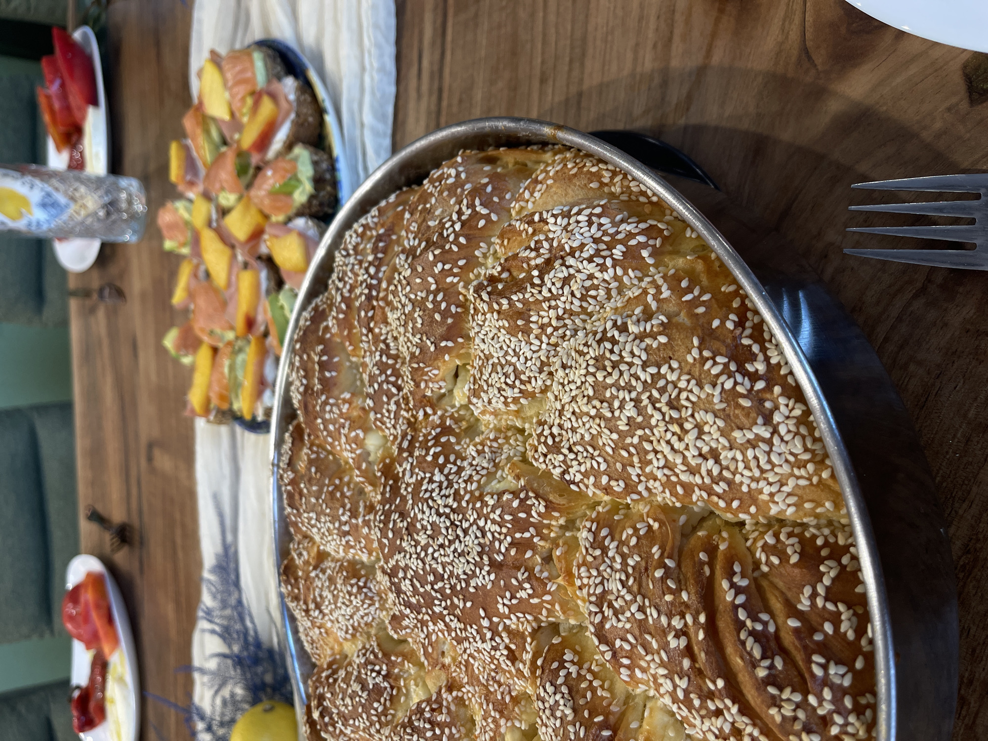 The image shows a close-up view of a round, golden-brown baked good generously sprinkled with sesame seeds, resting in a metal pan. The baked good appears to be divided into several interconnected sections, suggesting it might be a type of braided bread or pastry. The texture of the crust is visible, with some areas appearing slightly crispier than others.
In the background, on a wooden table, are other food items. To the left, a white plate holds pieces of red peppers and what looks like a white spread, possibly cheese. Further back, another plate displays small appetizers, appearing to be open-faced sandwiches or canapés topped with salmon, avocado, and mango. A glass with a yellow liquid and a lemon are also visible on the table. The setting seems to be a dining table, with hints of upholstered furniture in the blurred background.
The scene is set in the city of Karlovo, Bulgaria, though no specific landmarks or indications of this location are visible within the image itself. The lighting suggests it is daytime, possibly indoors or on a shaded patio. No people are present in the frame.