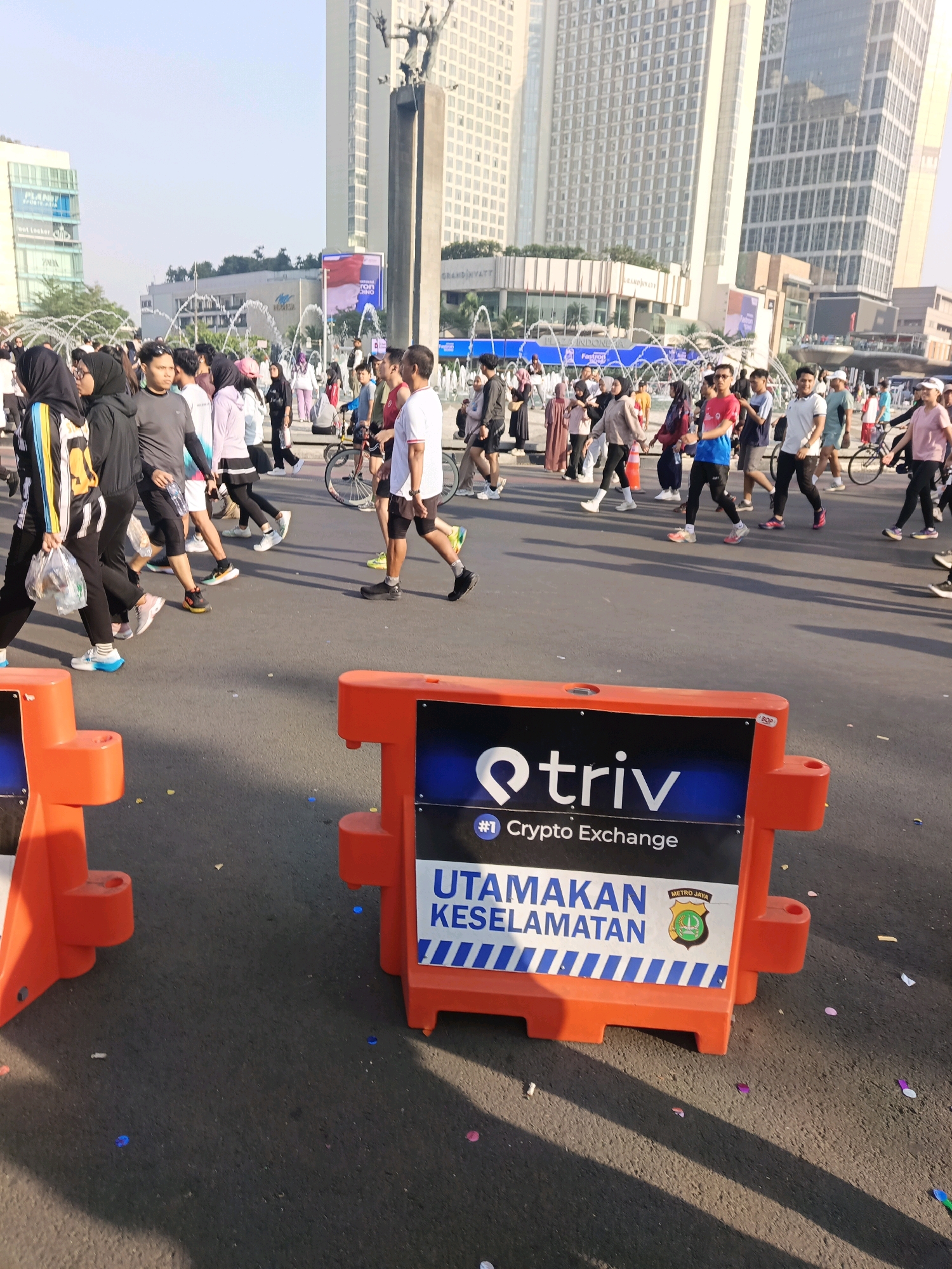 This image captures a busy street scene in Central Jakarta, Indonesia, likely during an outdoor event or a marathon given the presence of many pedestrians, cyclists, and the general atmosphere of a public gathering. In the foreground, prominent orange traffic barriers with signage are placed on the asphalt road, one displaying a "Triv" logo for a crypto exchange and the words "UTAMAKAN KESELAMATAN" (Prioritize Safety) along with a Metro Jaya police emblem. A crowd of people, predominantly dressed in casual sportswear and some in hijabs, are walking on the road, suggesting it's a pedestrianized area for the event. The background reveals the urban landscape of Central Jakarta, with modern high-rise buildings, including what appears to be the Grand Hyatt hotel, towering over the scene. Fountains are visible in the middle ground, adding to the urban setting. The lighting suggests it is daytime, with shadows cast on the road by the people and objects. The presence of confetti on the ground indicates a festive or celebratory element to the event.