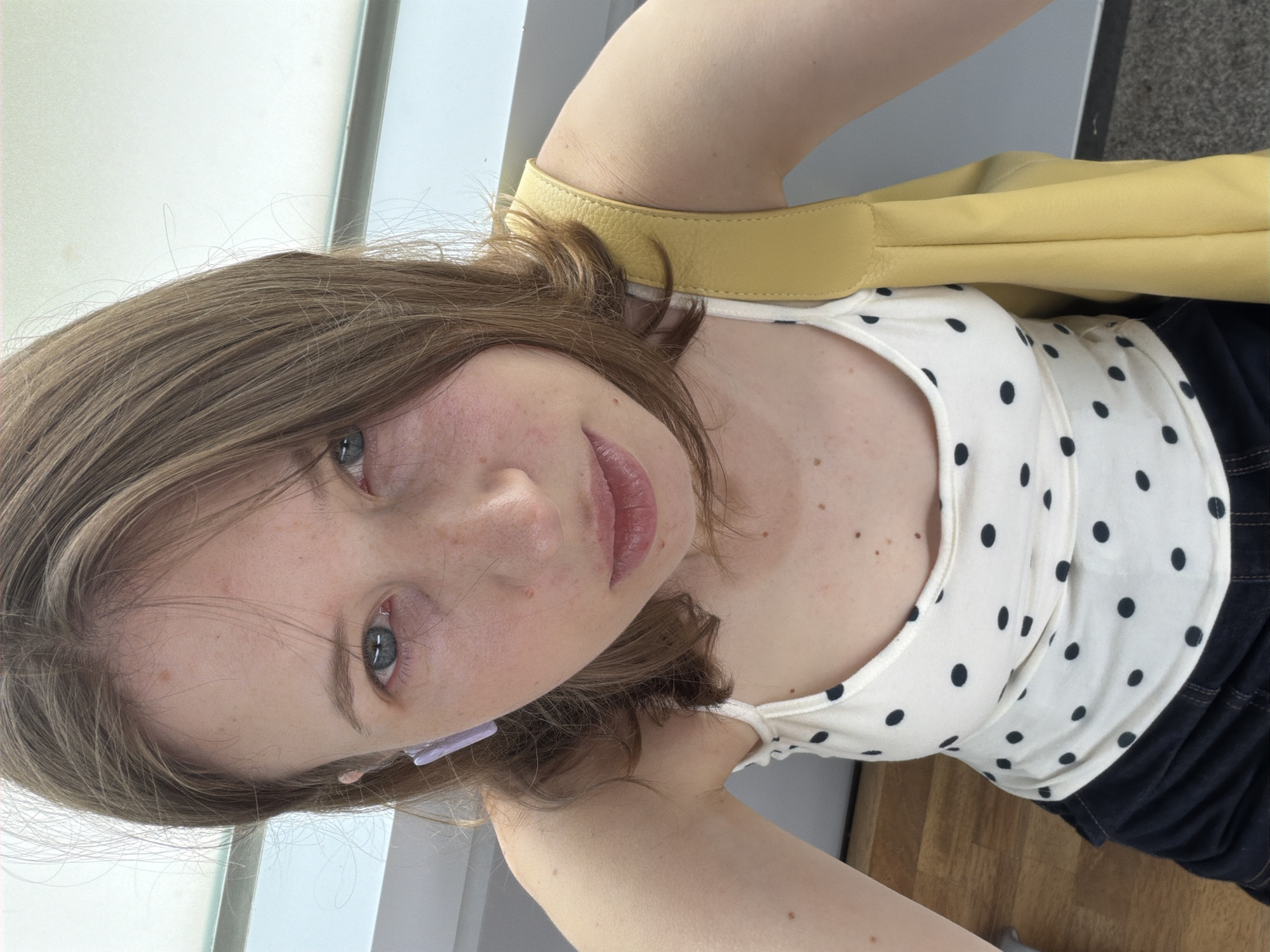 A young woman is taking a selfie in an indoor location. She has fair skin with freckles, light brown hair, and blue eyes. She is wearing a white tank top with black polka dots, dark shorts, and a light purple earbud. A yellow bag is slung over her shoulder. The background includes a wooden floor and a gray wall.