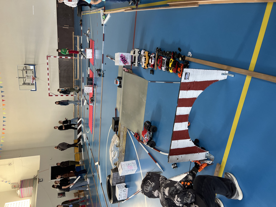 The image depicts an indoor remote-controlled (RC) car event taking place within a school gymnasium in Budva, Montenegro. The gymnasium features a bright blue sports court with yellow and white demarcation lines, high white walls, and a light-colored ceiling decorated with colorful triangular bunting. A basketball hoop with a clear backboard and a red rim, along with a handball goal with a white frame and black netting, are visible on the right side of the space.
A temporary RC car track is laid out on the court, constructed from various elements. These include a large grey metal ramp, multiple white cardboard boxes resembling buildings (one with "BAY!" graffiti), a black tire, wooden planks defining track edges, and a prominent red and white striped archway serving as a gate or checkpoint. Two rectangular red mats are positioned in the middle ground, and smaller red and white striped barriers delineate parts of the track.
Approximately 15-20 individuals, a mix of adults and younger people, are present, predominantly standing around the perimeter of the track, observing the activity. In the foreground, a person wearing a black and white patterned hoodie, dark pants, and grey sneakers is kneeling, actively operating an orange RC controller. This individual is positioned near a black and red monster truck-style RC vehicle, which is on the track near the red and white archway. A light blue and white monster truck RC car is visible on the grey ramp, and a smaller RC vehicle is behind the archway. A line of 10-12 parked RC vehicles, comprising various models including vans, cars, and trucks, is arranged beside the track near the archway. Some loose RC car parts are scattered on the track surface.