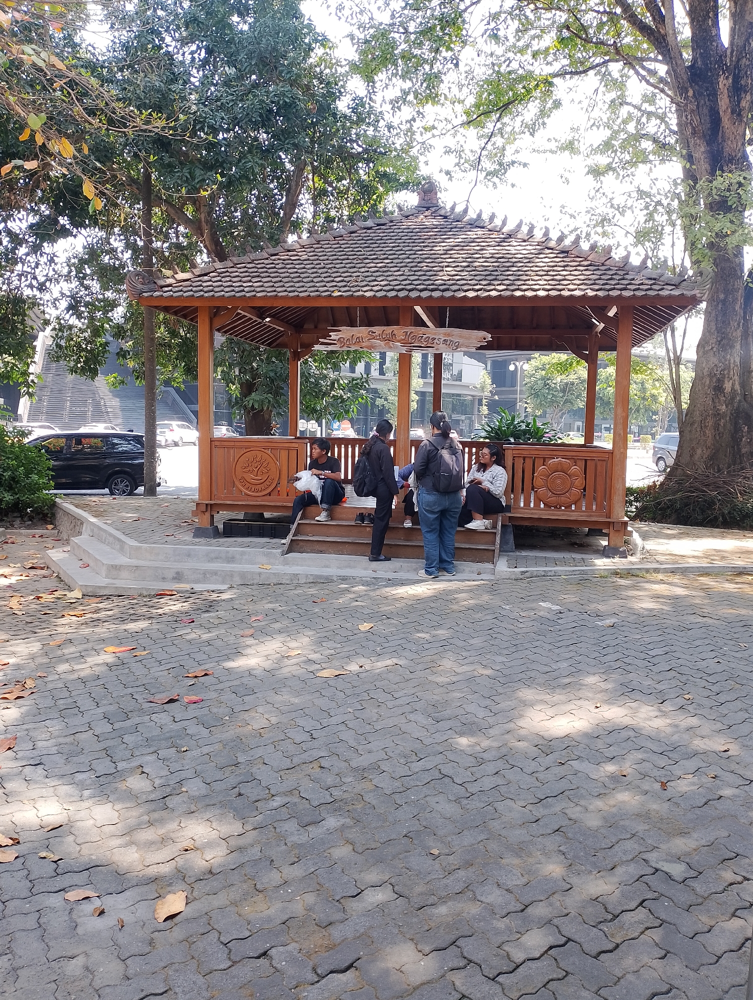 The image shows an outdoor gazebo in Depok, Indonesia, with four young people seated inside. The gazebo is made of wood and has a traditional Indonesian roof design. The ground is paved with interlocking concrete blocks, and fallen leaves are scattered across the surface. In the background, there is a large tree, a building with a modern facade, and a black SUV parked on the left. One person is holding a plastic bag, and another is looking at their phone. The overall scene suggests a casual gathering in a public space. The sign above the gazebo reads "Balai Sukajaya".