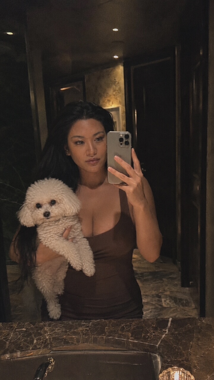 A woman with long, dark hair takes a mirror selfie while holding a small, white poodle. She is wearing a brown, sleeveless top and is in a dimly lit room with marble countertops and dark wood accents. The background appears to be a bathroom or a dressing area. The image does not contain any information about the city of Moscow, Russia.