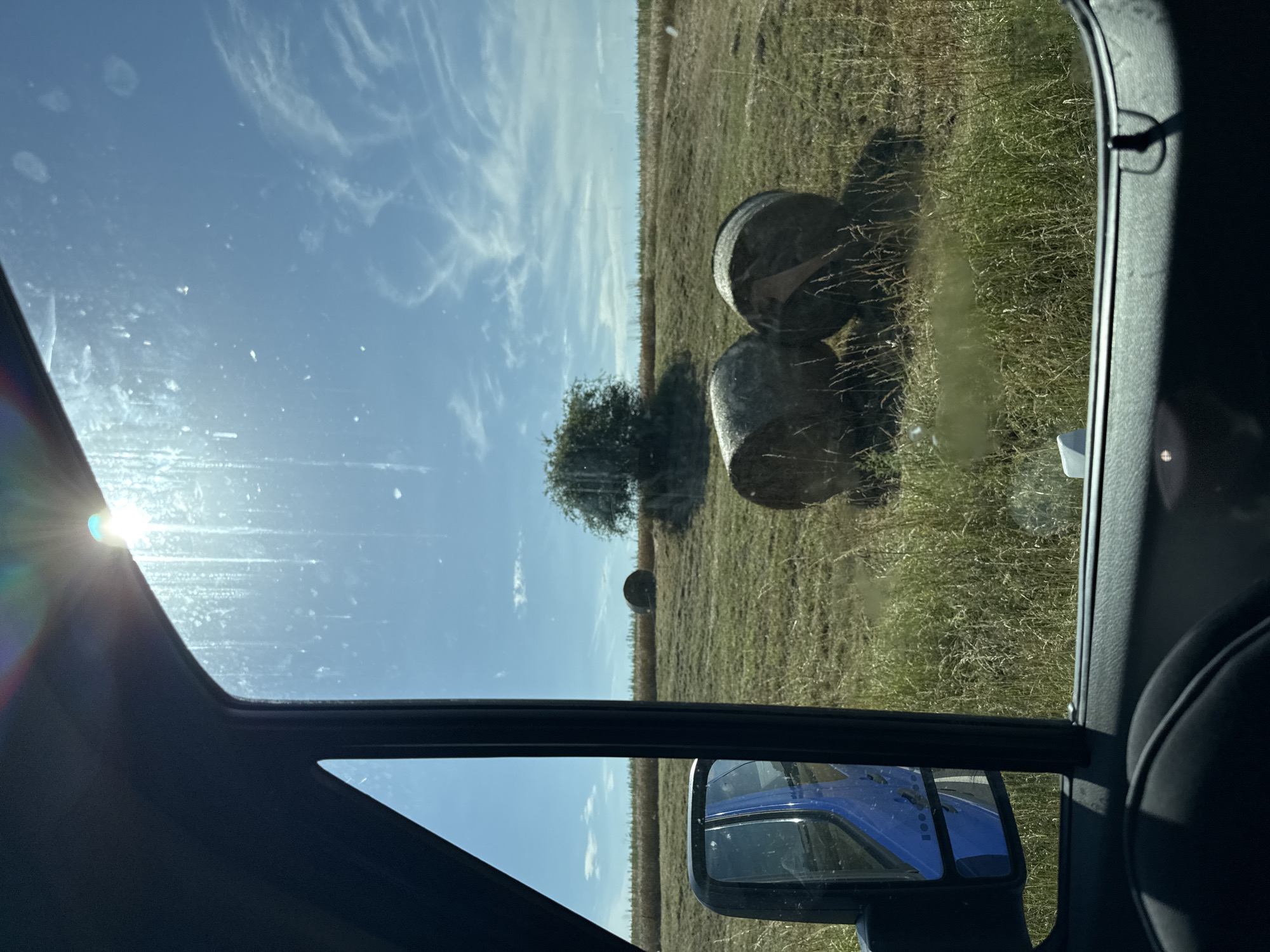 The image depicts a view from inside a vehicle, looking out at a rural landscape. The scene is dominated by a field of dry grass, likely hay, dotted with several large, round bales of hay. Two bales are prominently visible in the foreground, while another is further back in the field. A single, sparse tree stands in the middle ground, and a distant line of trees marks the horizon. The sky is a clear, bright blue with wispy white clouds, and the sun is shining brightly, causing lens flare in the upper left of the image. The presence of a side-view mirror reflecting a blue vehicle suggests the photo was taken while the vehicle was in motion or parked on a road adjacent to the field. The time of day appears to be late morning or early afternoon, given the angle of the sun and the brightness of the light. The overall impression is one of a rural, agricultural setting under a clear, sunny sky. No people are visible, and there is no discernible text in the image.
