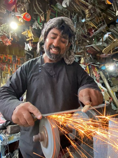 The image depicts a man in Kandahar, Afghanistan, sharpening a tool with a bench grinder. He is smiling and wearing a head covering and a dark, buttoned tunic. Sparks emanate from the point of contact between the tool and the grinding wheel. The background features a cluttered shop environment with various tools and metal objects hanging or stored overhead.