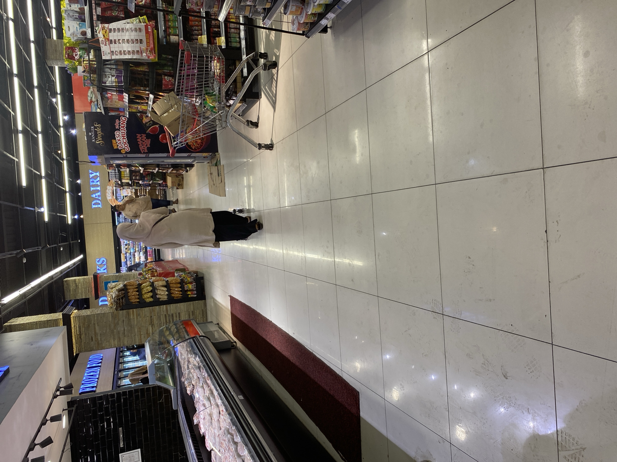 The image depicts an interior view of a grocery store, likely in Banda Aceh, Indonesia, given the context provided. The scene is characterized by bright overhead lighting illuminating tiled floors and aisles of merchandise. In the foreground, a person wearing a light-colored hijab and a long, flowing garment walks away from the camera, pushing a shopping cart. The shopping cart contains several items, including a cardboard box. To the left, a refrigerated display case filled with packaged food items is visible, with a dark red mat extending along its base. Further back, signage indicates sections such as "FROZEN FOOD" and "DAIRY." Shelves stocked with various packaged goods line the right side of the aisle. The floor shows scuff marks and footprints. The environment appears to be a typical supermarket setting.