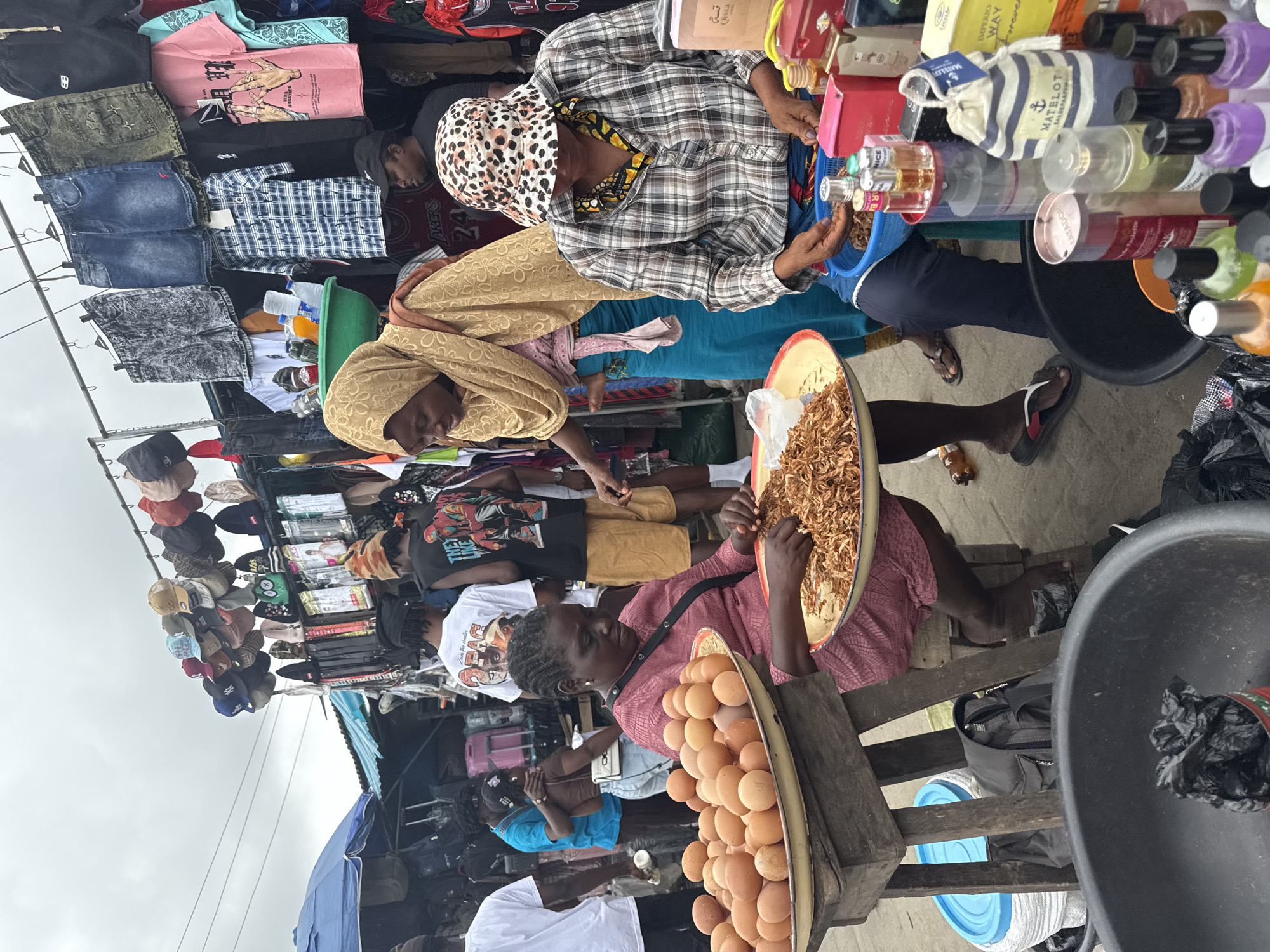 In Lekki, Nigeria, a bustling market scene unfolds with an abundance of goods and people. In the foreground, a woman in a pink dress is seated, holding a shallow dish filled with dried food items, possibly shrimp or fish, which she is sifting through. Beside her, a large tray overflows with brown eggs, illuminated by the overcast sky. To her right, another woman, wearing a leopard-print bucket hat and a plaid shirt, is actively engaged in sorting through a blue basin containing similar dried food. She is surrounded by an array of bottles, some appearing to be perfumes or oils, displayed in pink boxes. Behind them, a woman in a golden hijab and a blue skirt stands, also looking towards the food. The background reveals a vibrant market with stalls displaying clothing, including denim shorts, t-shirts, and hats. Several individuals are seen browsing or interacting within the market space. The environment suggests a typical day at an outdoor marketplace, with goods laid out for sale and shoppers engaged in commerce.