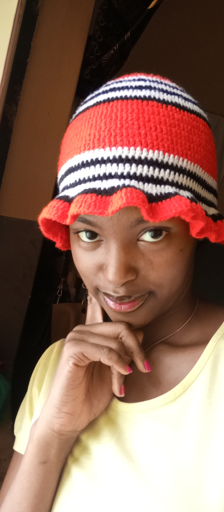A young woman is captured in a close-up, head-and-shoulders portrait, looking directly at the camera with a subtle, pleasant smile. She appears to be of African descent.
She is wearing a striking crocheted bucket hat that features a pattern of red, white, and black horizontal stripes. The brim of the hat is a solid red with a distinct ruffled edge. Her attire also includes a light yellow t-shirt, and a thin, delicate silver chain necklace is visible around her neck. Her fingernails are painted a vibrant pink. Partially visible behind her head on the right are dark earrings, possibly dangle style.
Her right hand is raised, with her fingers gently resting near her chin and lower lip, suggesting a thoughtful or posed gesture. Her eyes are open and engaging, and her lips have a glossy finish.
The setting appears to be indoors, with a warm, natural light illuminating her face. In the background on the left, a light brown wall or wooden frame is visible. On the upper right, a dark fabric is present, featuring a repeating pattern that resembles intertwined 'GG' shapes, characteristic of the Gucci brand logo. The overall ambiance is relaxed and inviting. No explicit, readable text is visible other than this background logo pattern.