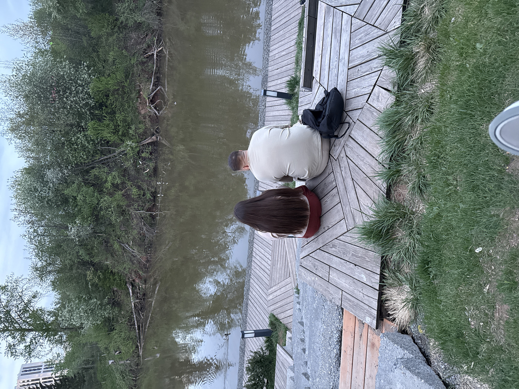 Two people, a man and a woman, are sitting on a wooden deck next to a body of water, potentially a pond or river. They are positioned with their backs to the viewer, looking out over the water. The man is wearing a light-colored shirt. The woman has long, dark hair and is wearing a dark shirt or dress. A black backpack sits next to the man.
The deck is made of wooden planks arranged in a geometric pattern. To the right of the deck is green grass, and to the left is the body of water, reflecting the surrounding trees and sky. Trees are along the bank of the water. Steps lead down from the deck towards the water. In the distance, a building can be partially seen.
The location is in Izhevsk, Russia.