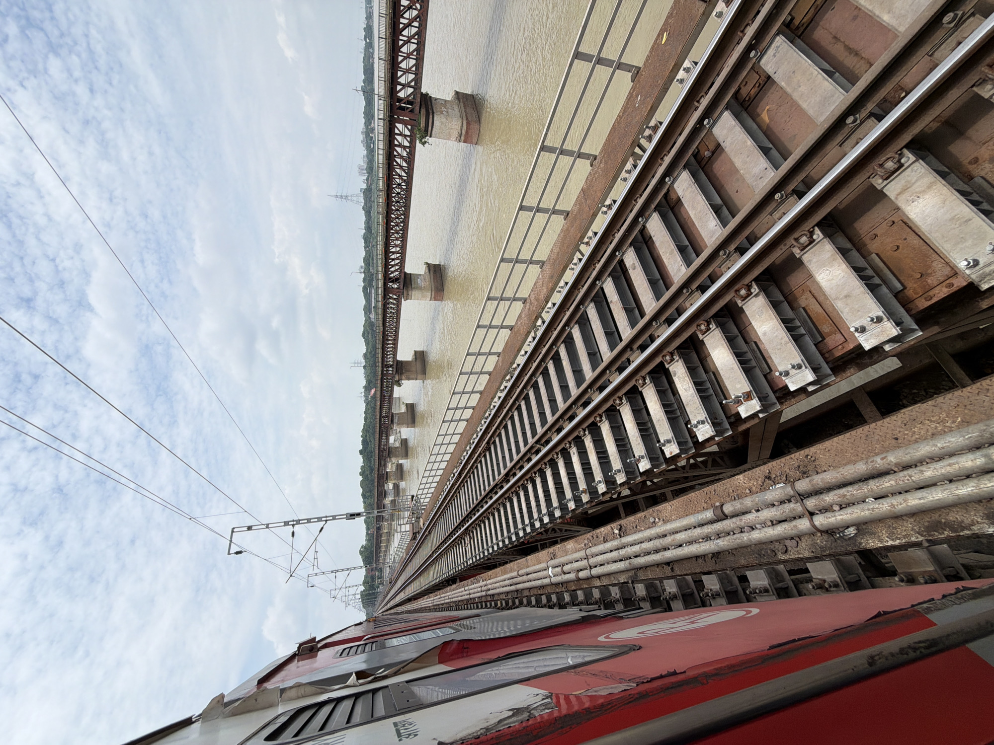 The image captures a view from a moving train on a bridge crossing a wide, murky river in Shuklaganj, India. The train, painted red and white, is on the left side of the frame, partially obscuring the view. The railway tracks stretch forward, disappearing into the distance. Parallel to the tracks is a metal railing. The river flows beneath the bridge, with a rusty, multi-span railway bridge visible in the background, also crossing the river. The sky is overcast with scattered clouds. The scene depicts a transportation infrastructure scene over a natural body of water.