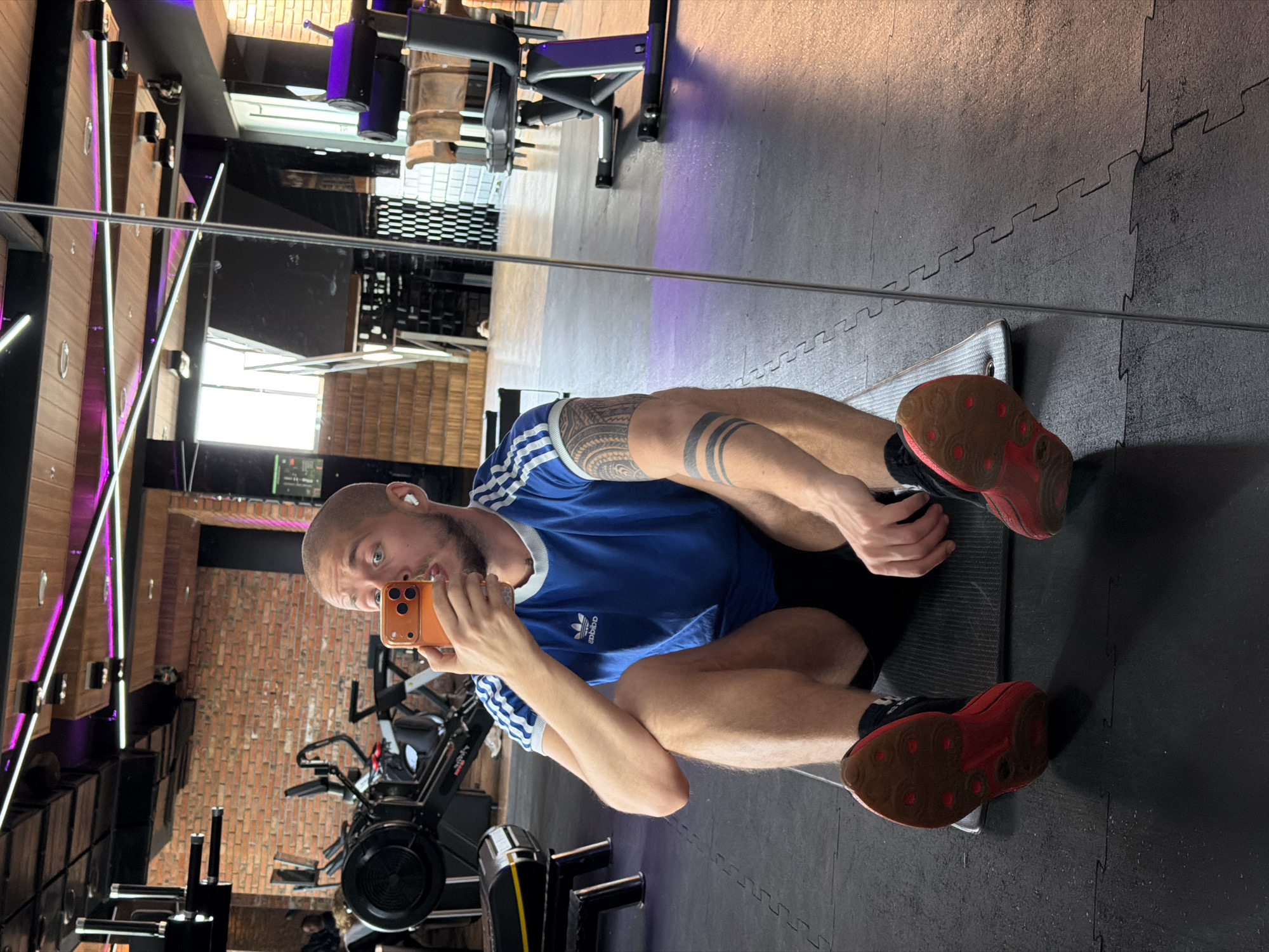 A man with a shaved head, light skin, and a beard is sitting on a mat in a gym, holding an orange smartphone to take a mirror selfie. He is wearing a blue Adidas t-shirt with white stripes on the sleeves and black shorts. He has tattoos on his left arm and thigh. His feet are bare, but he is wearing black socks and red athletic shoes. The gym appears to be a modern facility with exposed brick walls, wooden ceilings with purple lighting, and various exercise machines, including a rowing machine and weight machines. In the background, a staircase is visible, as is a large mirror reflecting the gym. The floor is covered with black puzzle mats. The image is taken in a gym in Sofia, Bulgaria.