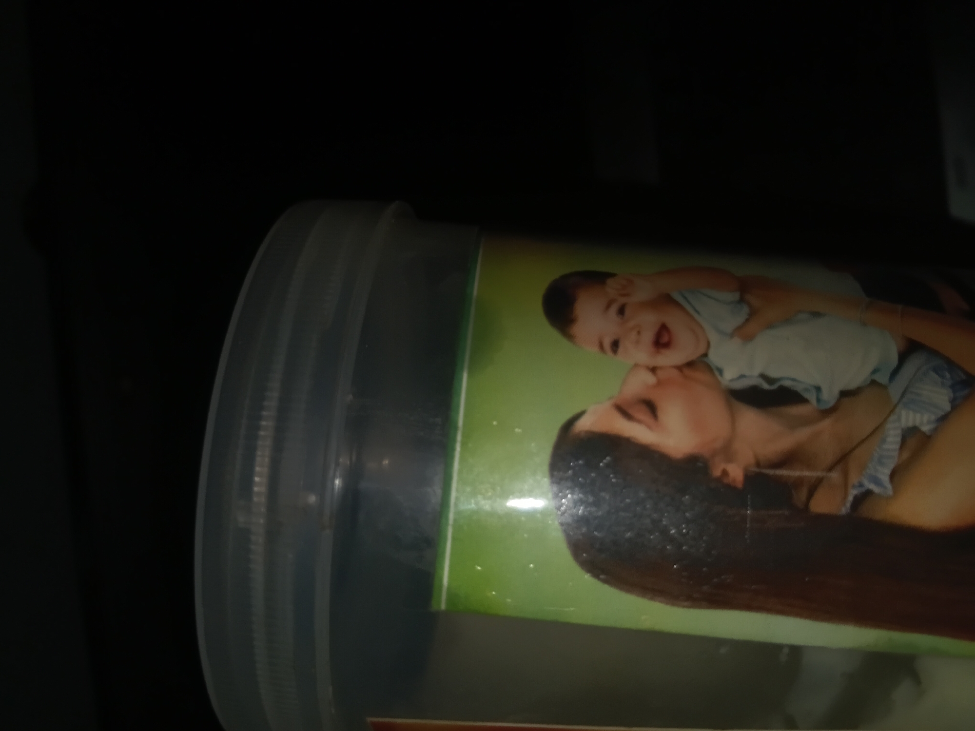 A woman with long brown hair is kissing a baby on a container with a green background and a picture of the two of them on it. The baby is smiling and has its arms up. The woman is wearing a blue and white striped top. The container has a clear plastic lid with ridges. The image is dark with some bright lights. The scene is likely in a home or shop in Jalingo, Nigeria.