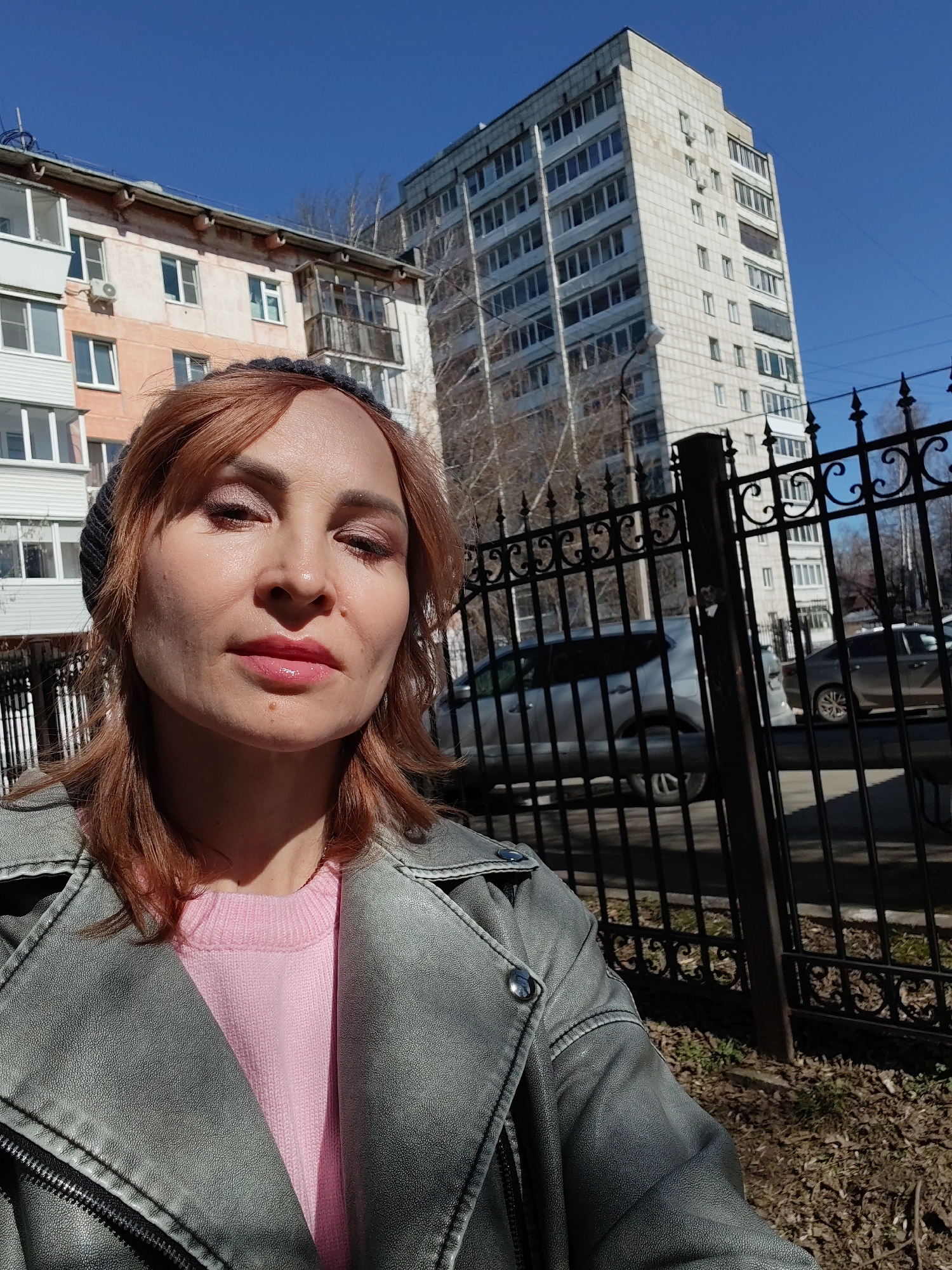 A woman is standing outside in Chaikovsky, Russia, on a sunny day. She is wearing a gray leather jacket over a pink sweater and a dark-colored knit hat. She has reddish-blonde hair and appears to be wearing makeup, including lipstick and eyeshadow. Behind her is a black metal fence, a parked car, and two multi-story apartment buildings. One building is a light peach color with white trim, and the other is a taller, white building. Some bare tree branches are visible. The ground near the fence is covered with dry leaves.