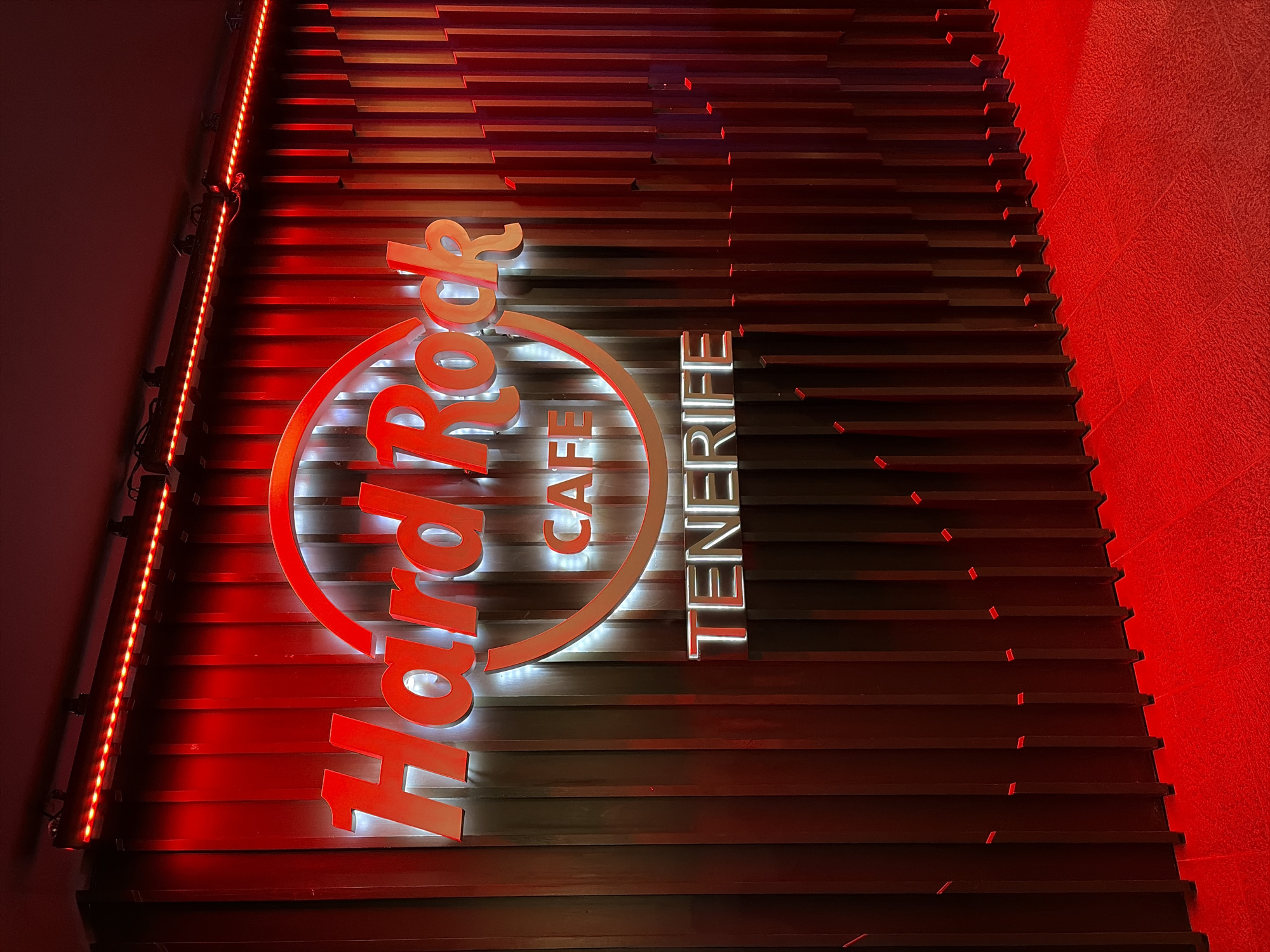 The image shows the illuminated Hard Rock Cafe Tenerife sign on a wall composed of horizontal slats. The location is implied to be in Los Cristianos, Spain. The sign is brightly lit, with red and white colors. The lighting creates a strong contrast between light and shadow on the textured wall. The overall impression is one of a modern, stylish, and well-lit establishment.