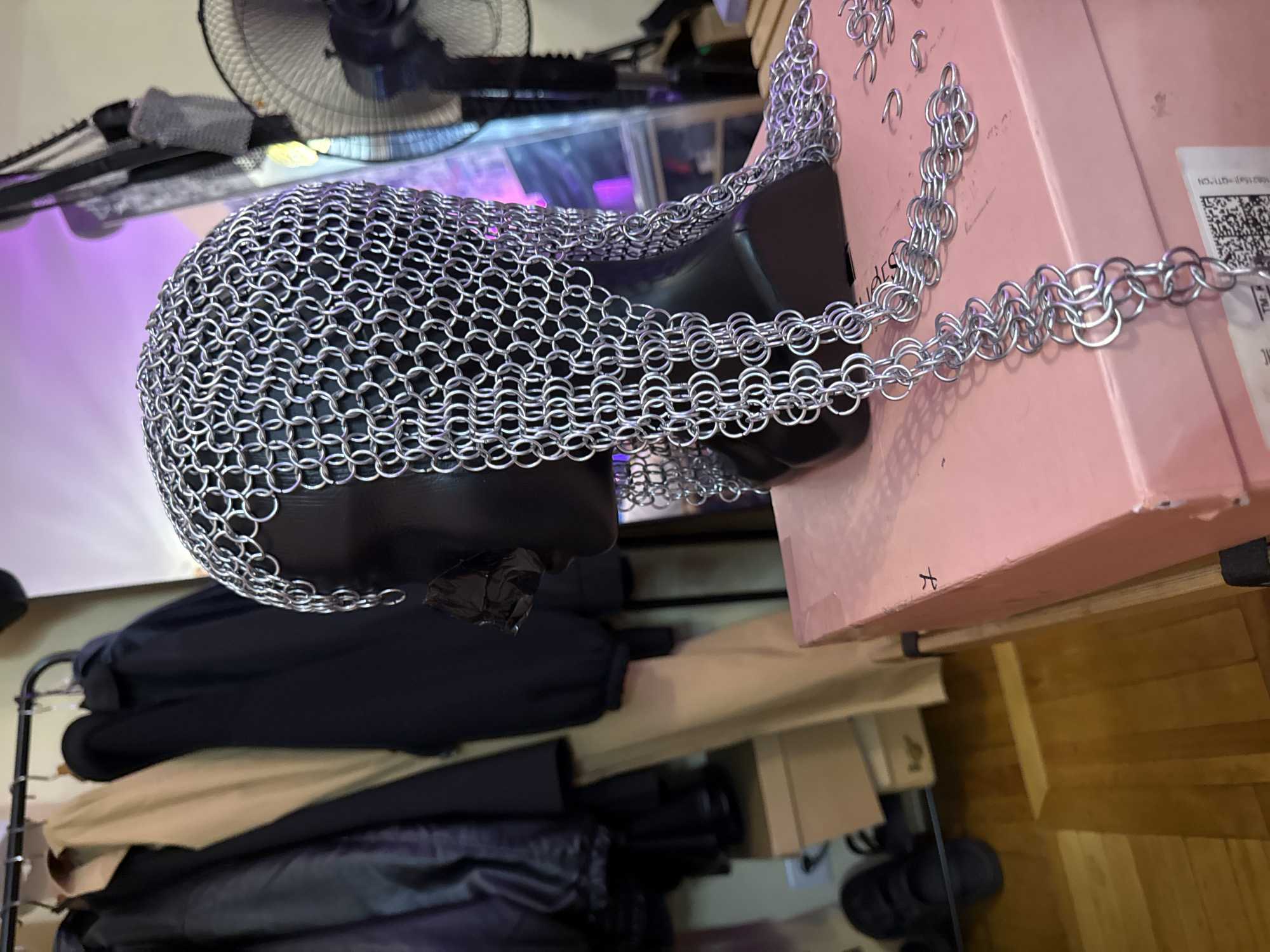 The image depicts a close-up view of a black mannequin head covered with a chainmail coif, resting on a pink box. The coif is intricate, made of numerous interlocking metal rings, and drapes down the side of the mannequin. A portion of a metal fan is visible to the right, and in the background, several coats are hanging on a rack. The scene appears to be indoors, possibly in a room with wooden flooring and cardboard boxes stacked behind the mannequin. There is no identifiable information that would suggest a location in Moscow, Russia, or any specific city.