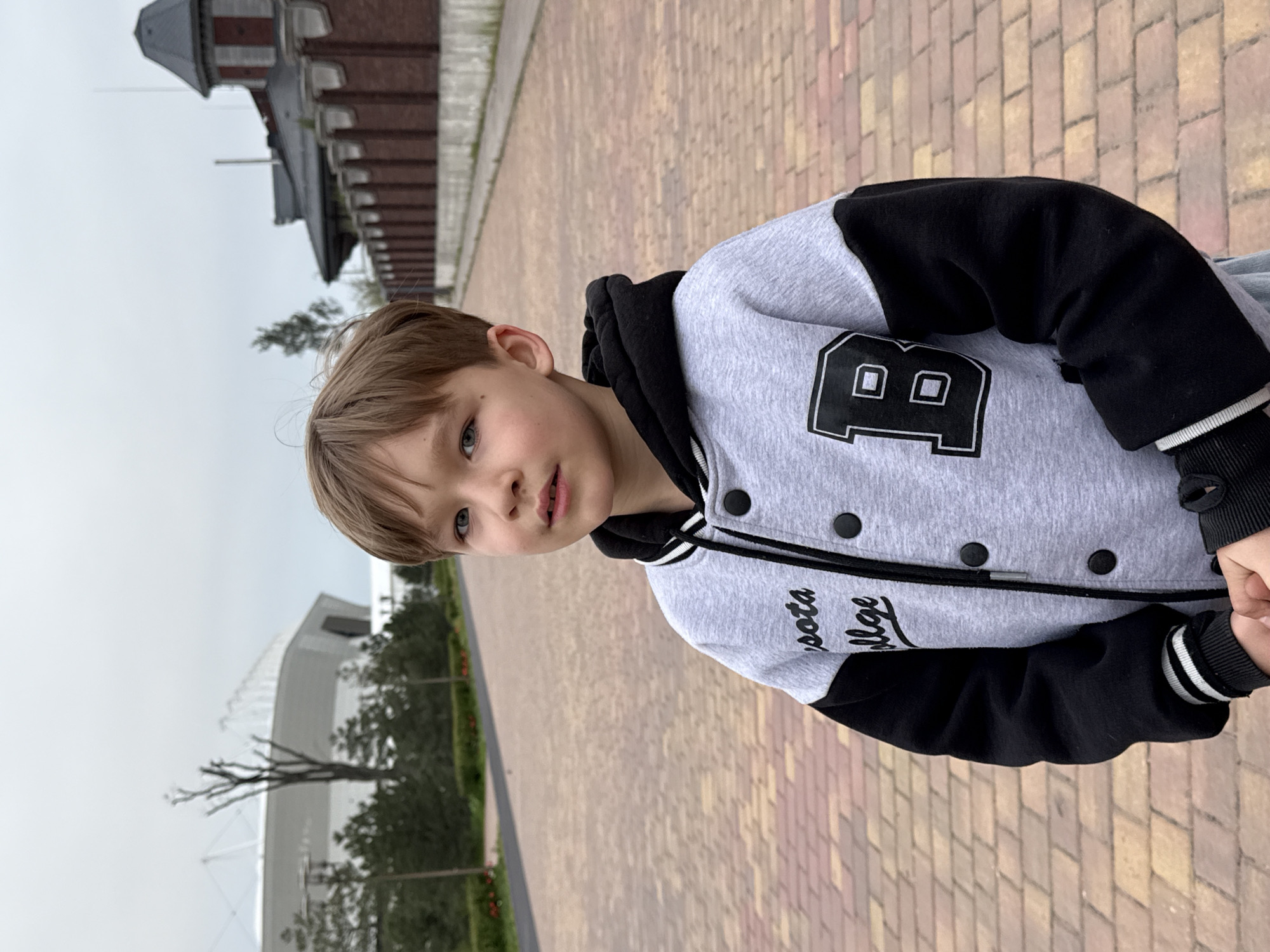 A boy is standing outdoors on a brick-paved walkway. He wears a grey and black varsity jacket with a large letter "B" and the word "College" written on it. In the background, a building with a distinctive tower structure is visible, along with greenery and what appears to be a modern stadium. The location is Rostov-on-Don, Russia.