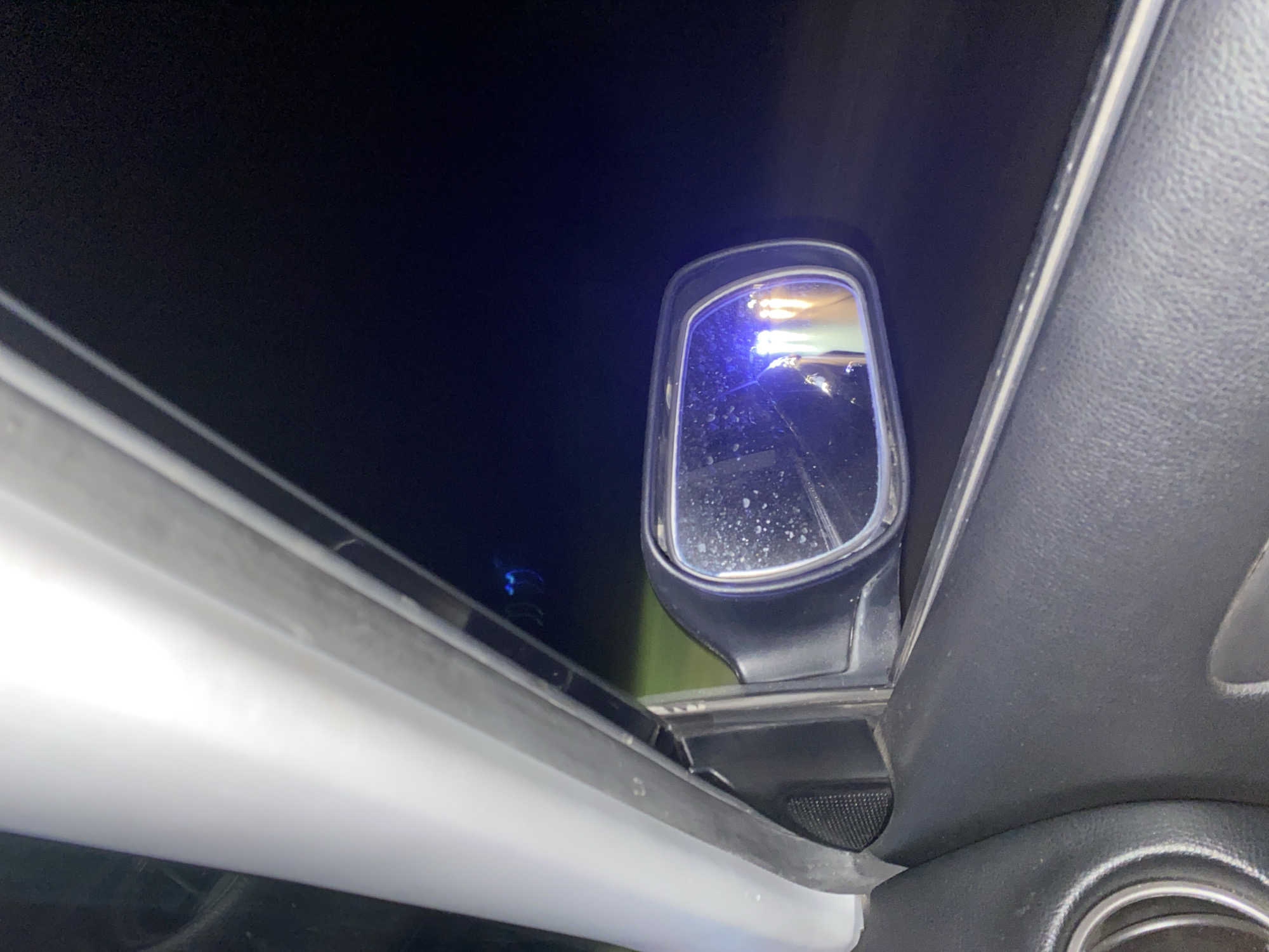 The image is taken from inside a vehicle, looking out the driver's side window at night. A side-view mirror is visible, reflecting lights that appear to be from another vehicle, possibly with flashing blue lights, suggesting a law enforcement presence. The interior of the car, including the door panel and speaker grille, is visible in the foreground. The background is dark, indicating nighttime. There are specks of what appear to be water droplets on the mirror. The location in the city of Yasen, Bulgaria is not determinable from the visual information provided.