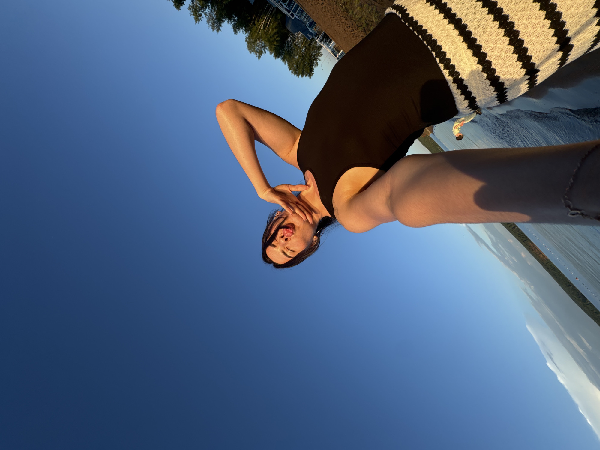 A woman is taking a selfie outdoors with a clear blue sky above. She is wearing a black tank top and a white and black striped skirt. Her tongue is sticking out and her hand is on her neck. She is standing near a body of water, possibly a lake or river, with a treeline and some buildings in the background. The sun is setting, casting a warm glow on her skin and the surrounding landscape. The location appears to be a waterfront area in Chaikovsky, Russia, characterized by a calm body of water, a sandy or dirt embankment, and vegetation typical of the region.