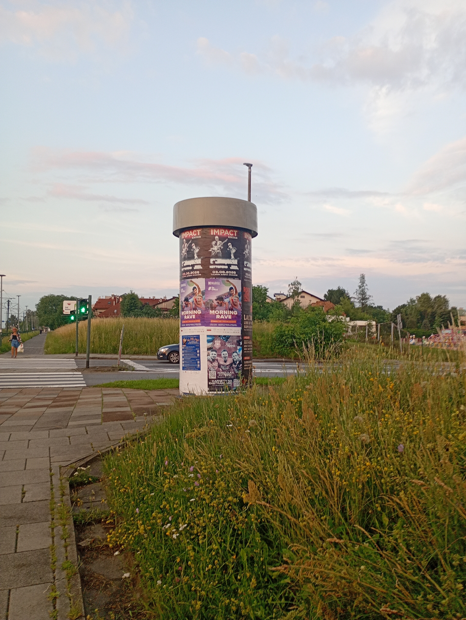 The image shows a cylindrical billboard with multiple posters on it, located in Krakow, Poland. The billboard is situated next to a pedestrian crossing and a road with a car driving on it. The scene appears to be in an urban environment with trees, buildings, and a grassy area with wildflowers in the background. The sky is visible with some clouds. A woman is walking on the sidewalk in the left part of the image. A traffic light is also visible.
