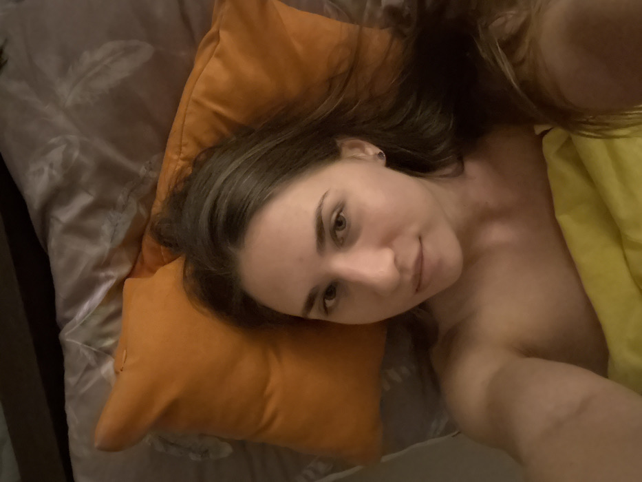 A woman with long brown hair lies in bed. Her head rests on an orange pillow. She gazes at the camera with a slight smile. A yellow blanket covers part of her body. The bedsheets are gray with a subtle feather pattern. Her skin appears smooth and clear. The lighting is soft and diffused, creating a warm, intimate atmosphere.
