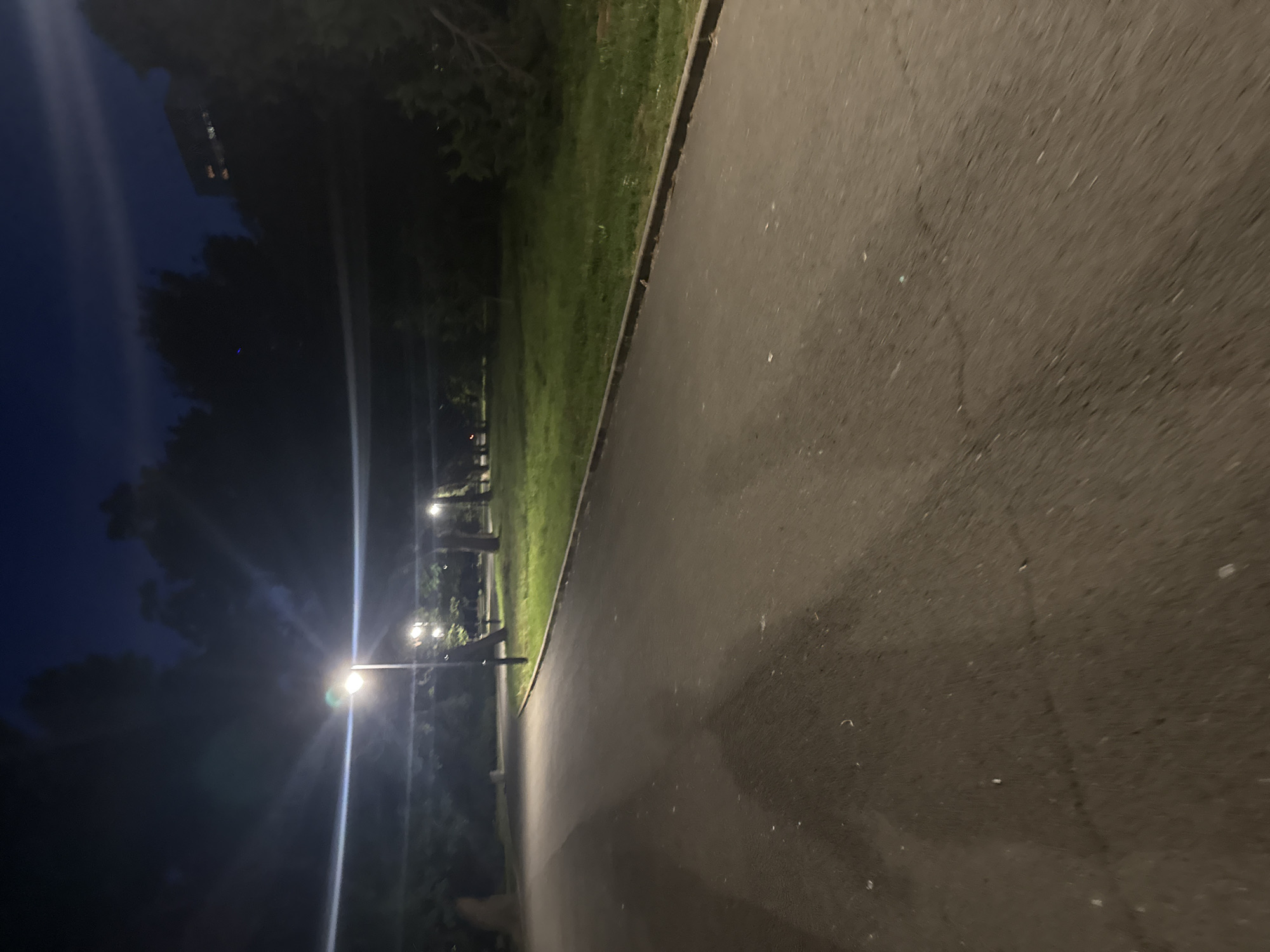 The image depicts an asphalt pathway at night, illuminated by lampposts, in a park area. The pathway curves to the left, with a grassy area and trees on the right. A building with illuminated windows is visible in the background, set against a dark night sky. The overall scene suggests a public park in an urban setting. Given that the location is Burgas, Bulgaria, this scene could be within the Sea Garden (Morska Gradina), a well-known park in the city that features pathways, green spaces, and views of the sea, though the sea is not visible in this particular frame. The lighting suggests the path is used at night, possibly for walking or jogging.