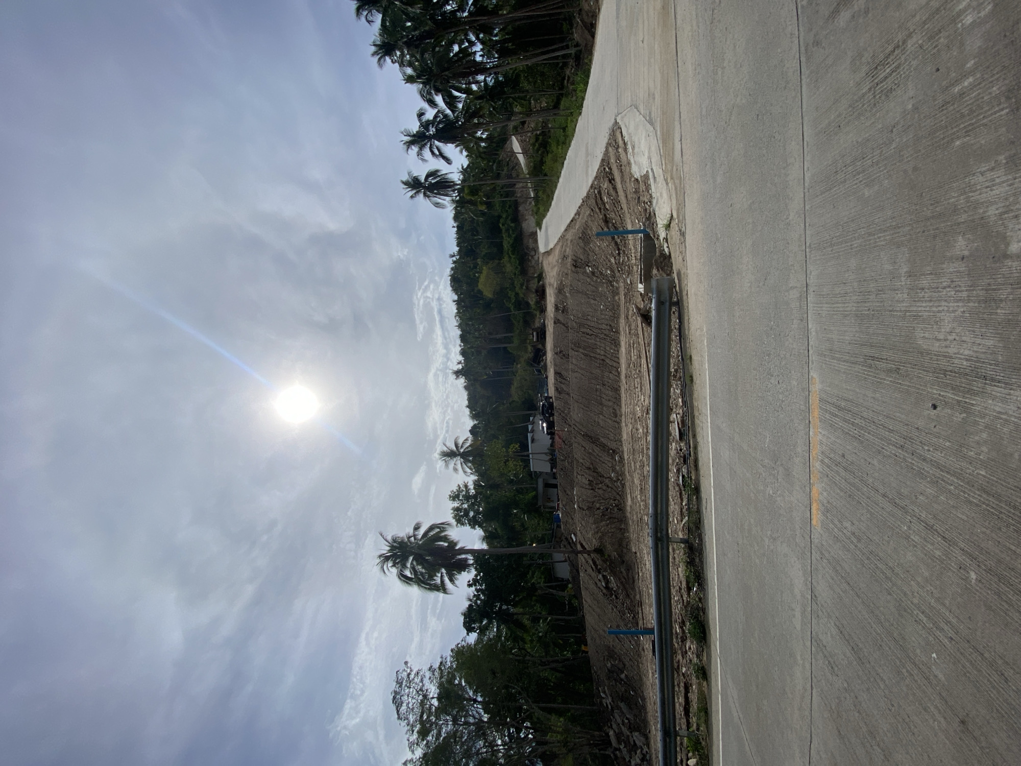 The image depicts a concrete road in Baan Wang Ta Kien, Thailand. The road curves along the edge of a hillside densely covered with trees, including palm trees. A rough, dirt shoulder separates the road from the hillside. A metal guardrail runs along the roadside. The sky is visible above the trees and is partly cloudy with the sun shining through. There are no visible people.