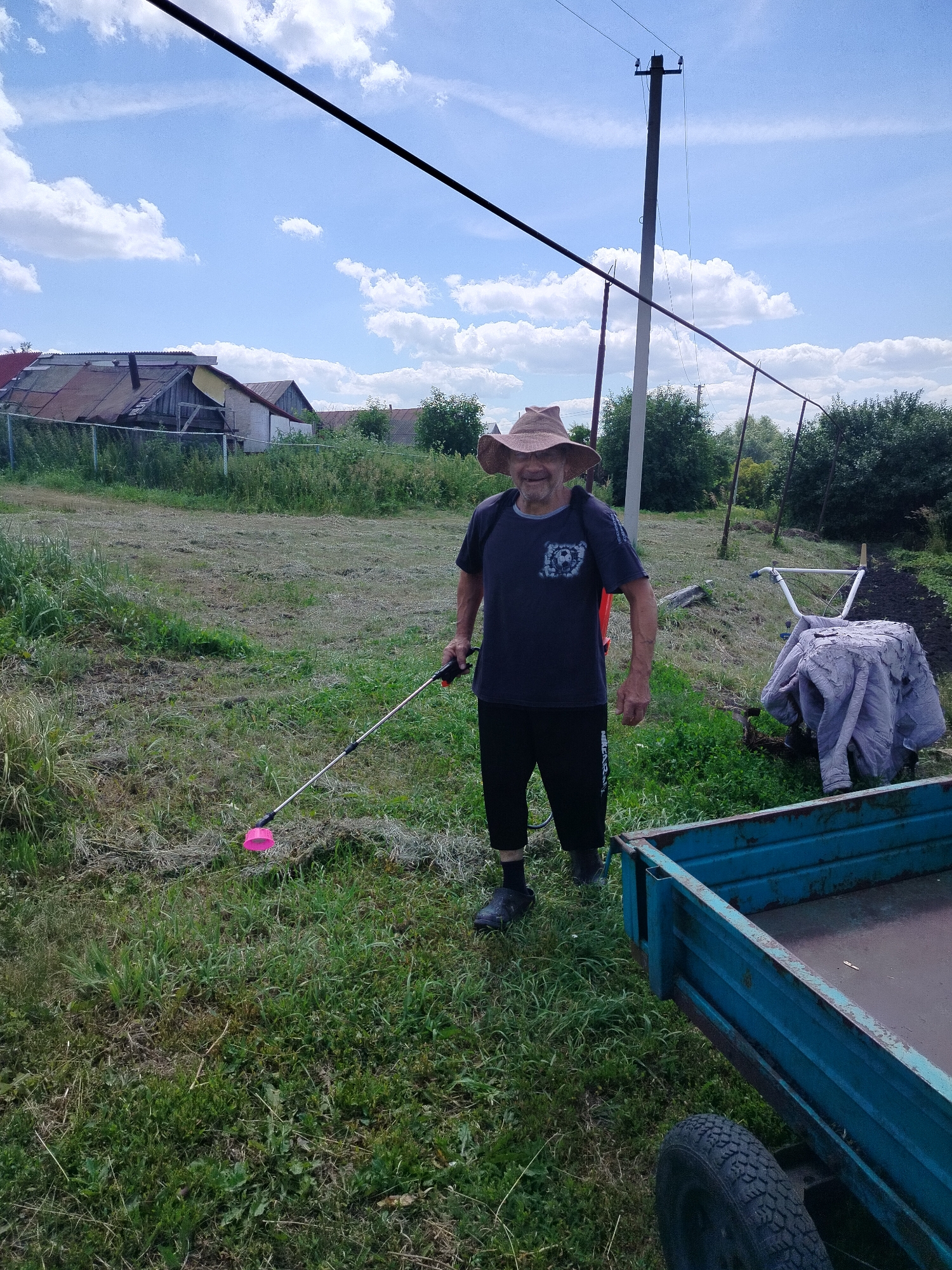 In an outdoor scene in Блохино, Russia, a man wearing a wide-brimmed hat and a dark t-shirt stands in a grassy yard. He is holding a long, thin pole with a pink object at its end, possibly a weed wacker or a similar gardening tool. The man is smiling and appears to be engaged in yard work.
Behind him, a power pole stands tall with wires extending across the frame. To his right, a garden tiller covered with a light-colored cloth is visible, along with a blue metal trailer. The background features houses with weathered roofs and dense green trees, suggesting a rural or semi-rural environment. The sky is bright blue with scattered white clouds, indicating a pleasant, possibly midday, time of day and fair weather. The overall impression is of a man working outdoors on a sunny day in a rustic setting.